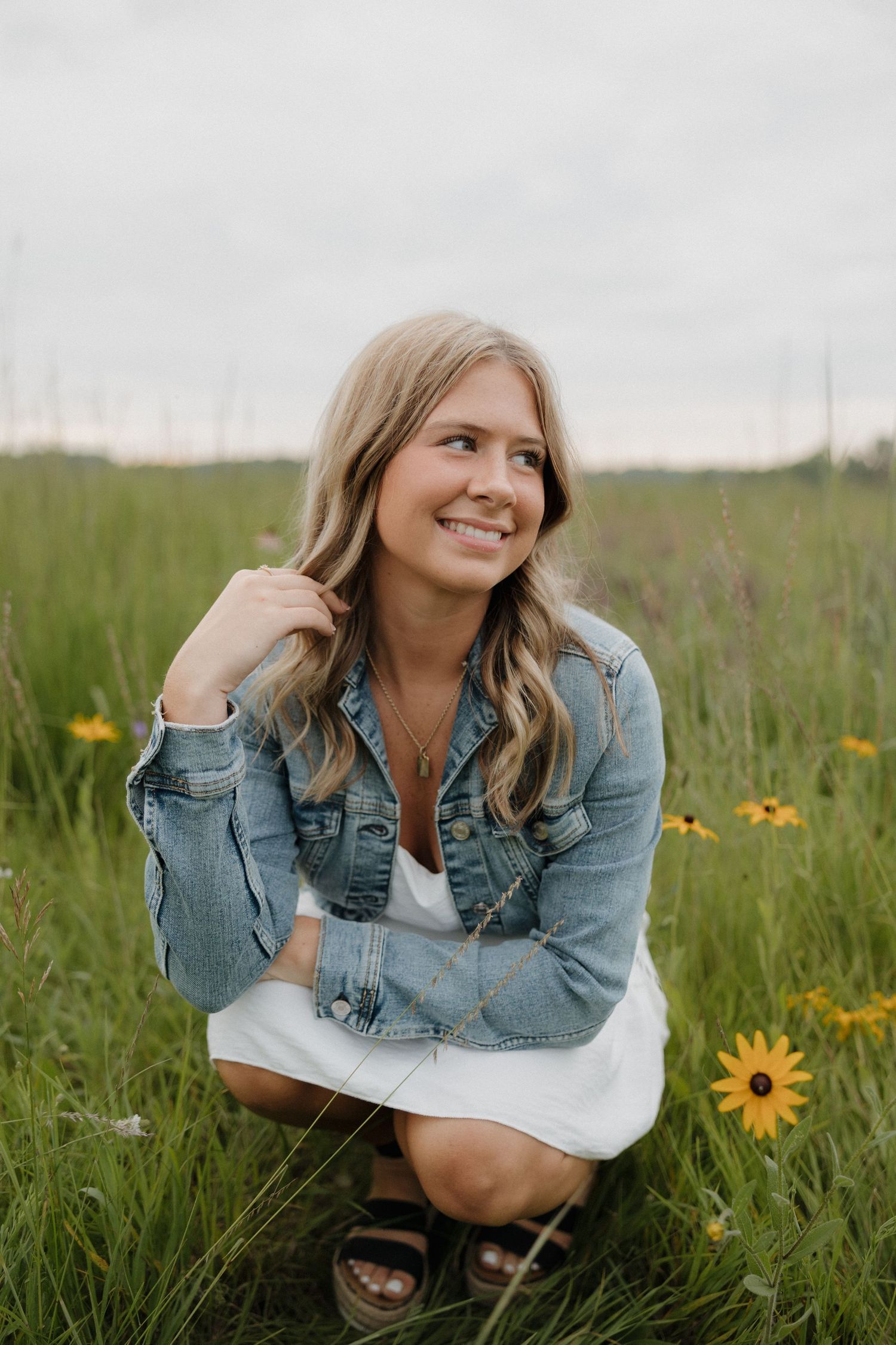 Blonde woman in denim jacket and white dress crouching among yellow wildflowers in a green field.