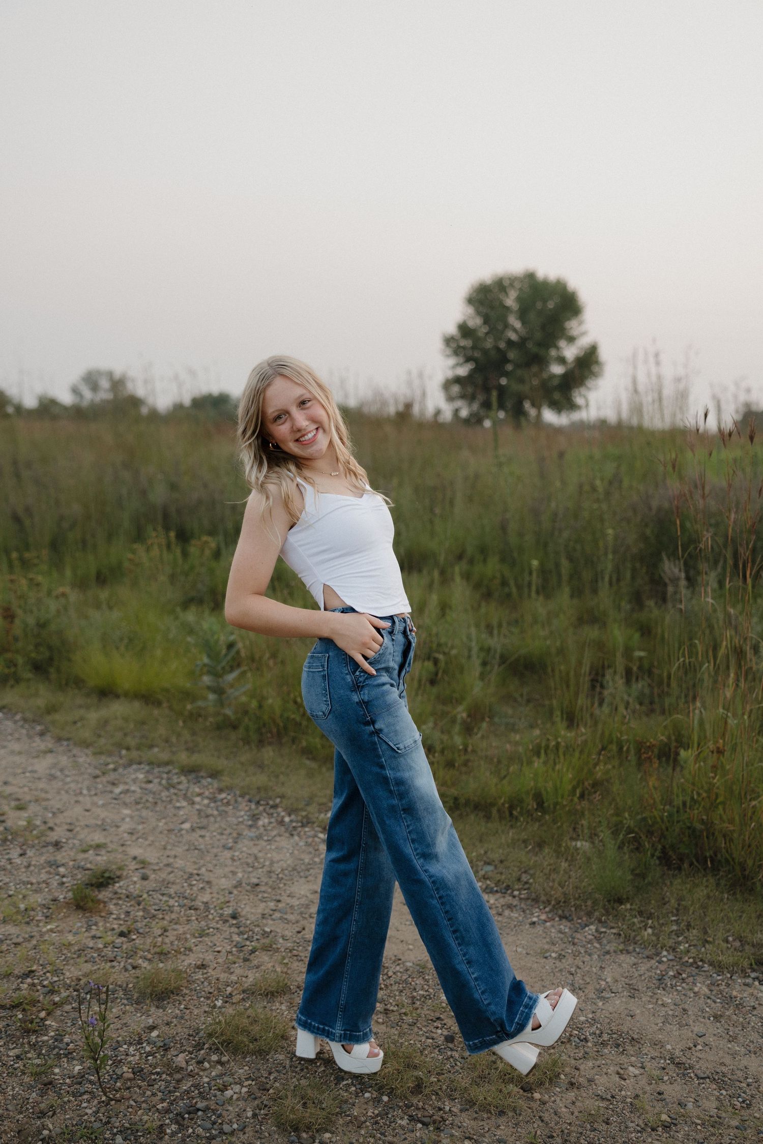 Blonde woman in white tank top and blue wide-leg jeans posing on gravel path surrounded by lush greenery.