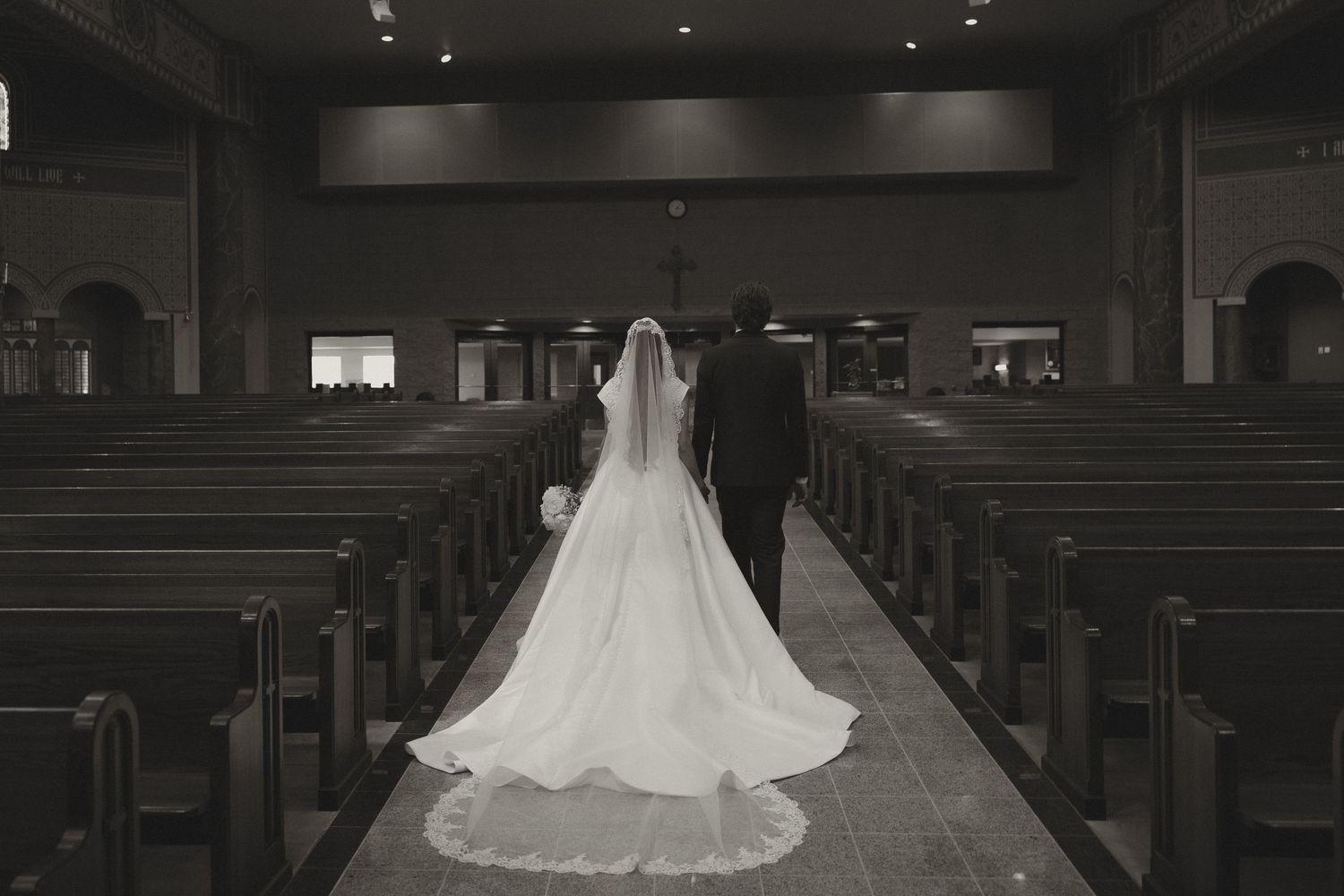 A bride in a flowing white wedding gown stands alone in the center aisle of a dimly lit church sanctuary.