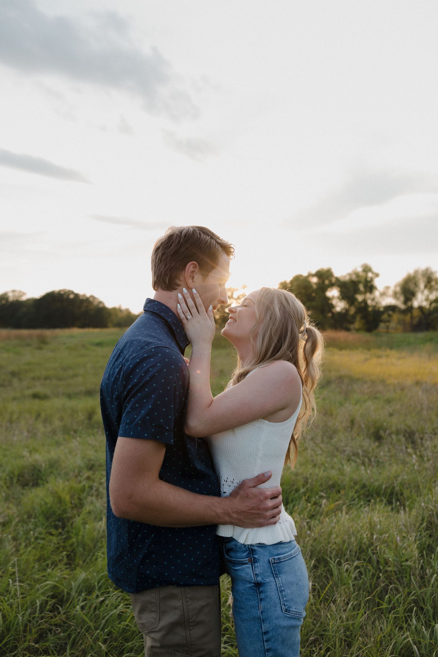 Couple sharing a tender kiss at sunset in a lush green field, woman touching mans face gently.