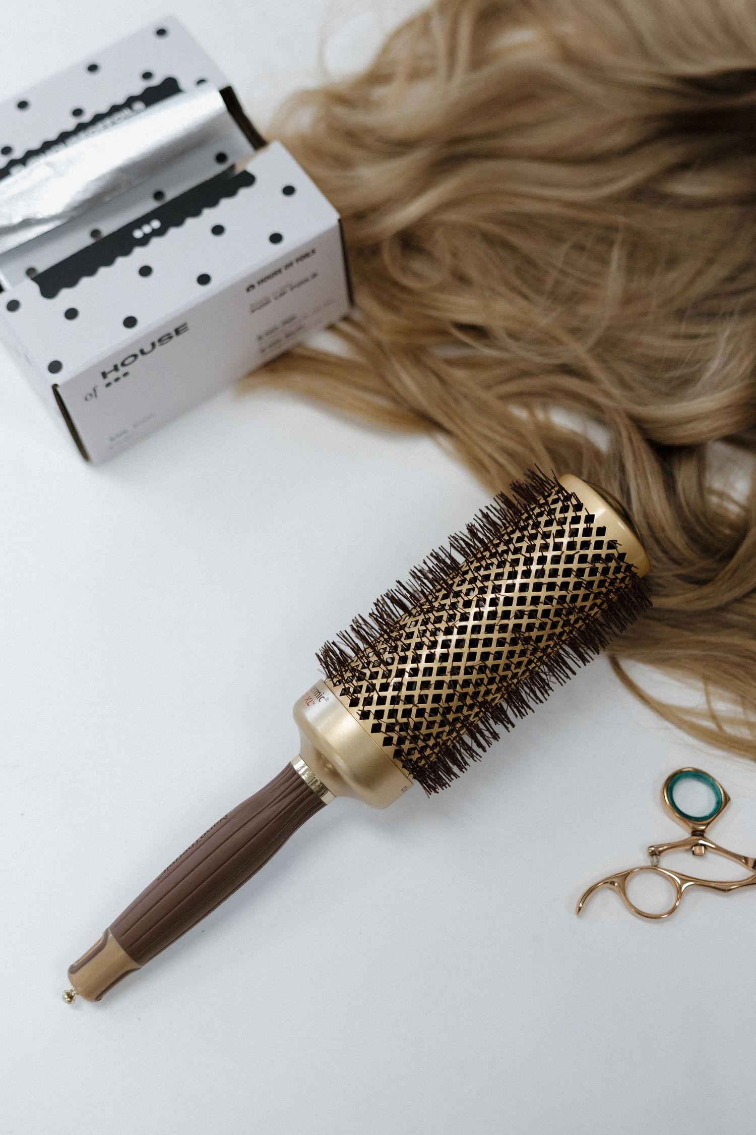 Gold barrel hair brush with wooden handle beside blonde wavy hair extensions and styling accessories on white surface.