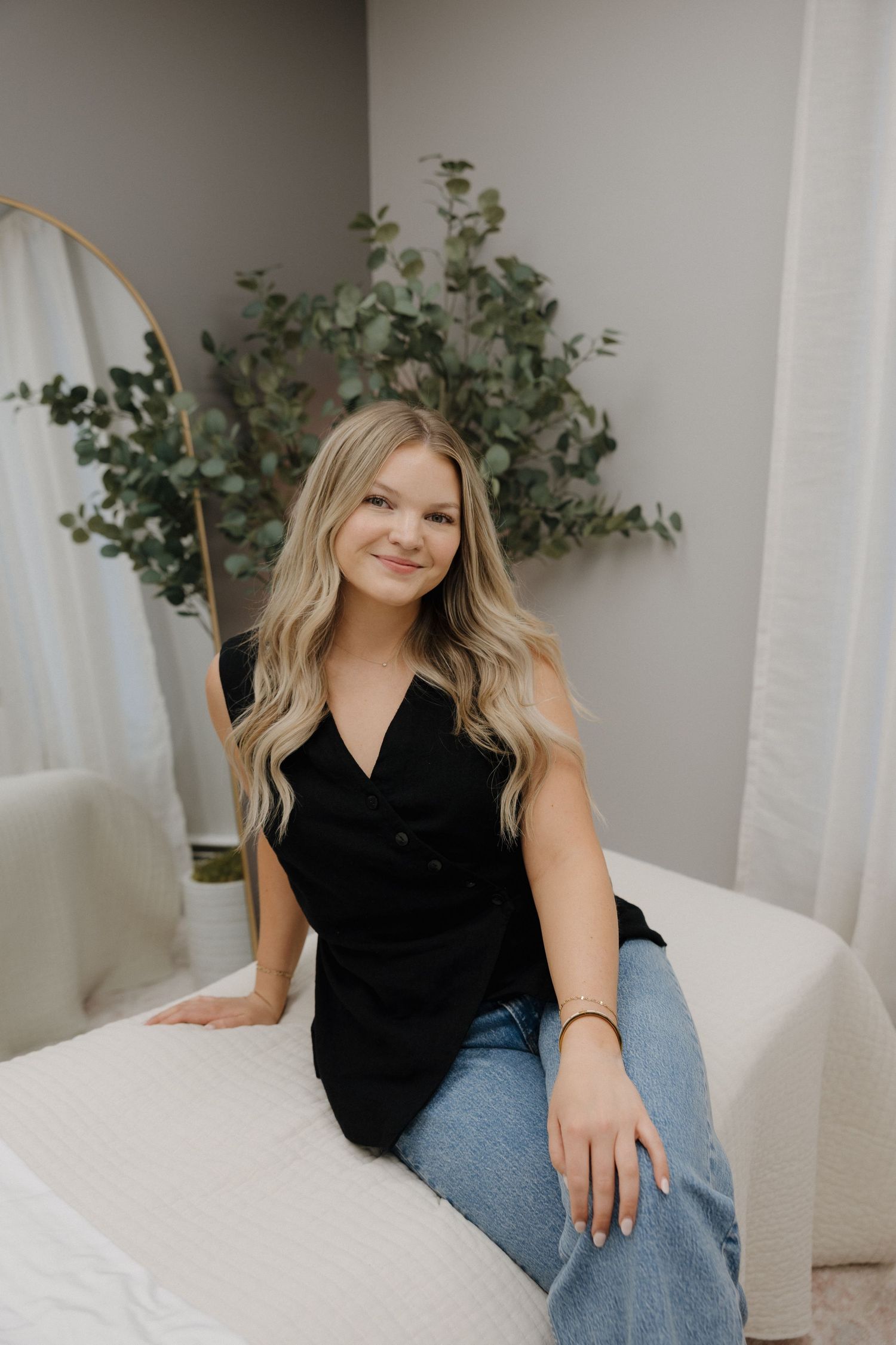 Blonde woman in black sleeveless top and jeans sitting on bed with eucalyptus decor behind her.