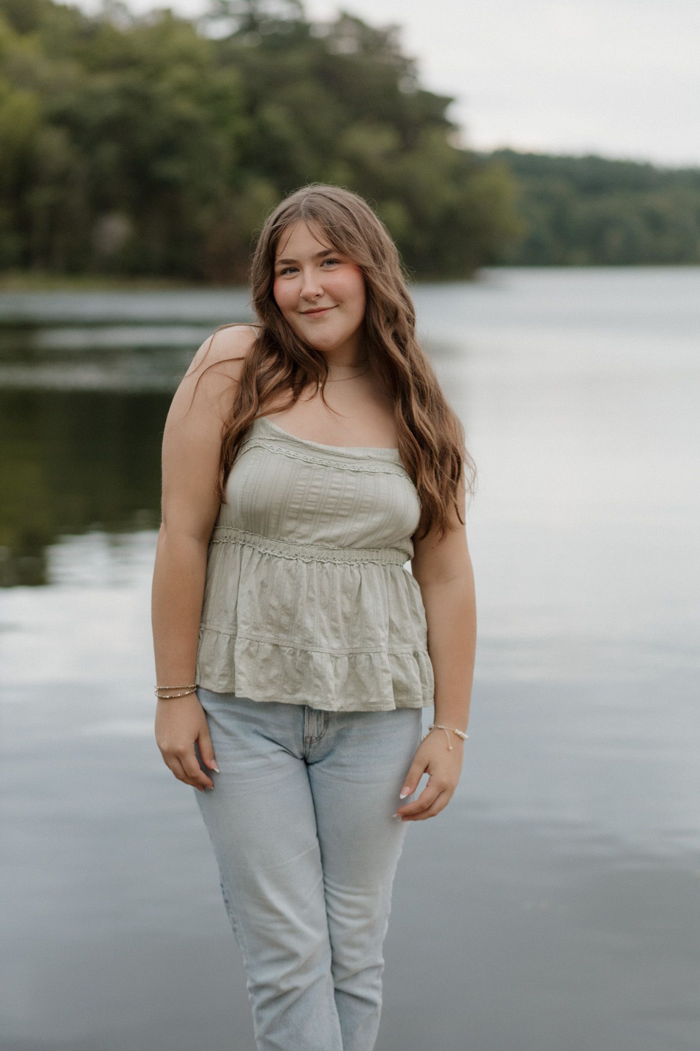 Smiling teen girl in sage green top and jeans standing near a peaceful lake shore.