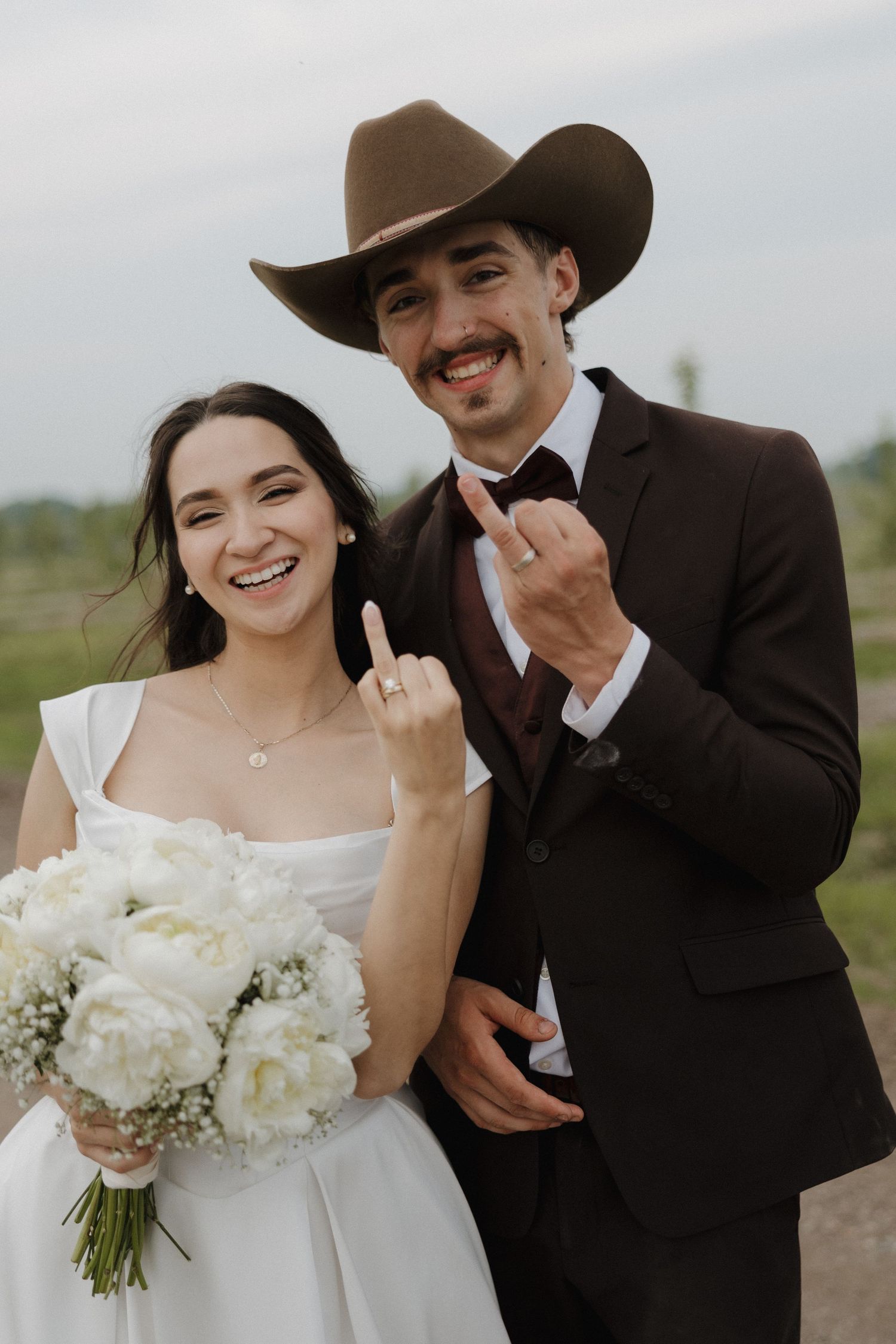 A couple shares joyful moments together in their wedding attire against a cloudy outdoor backdrop.
