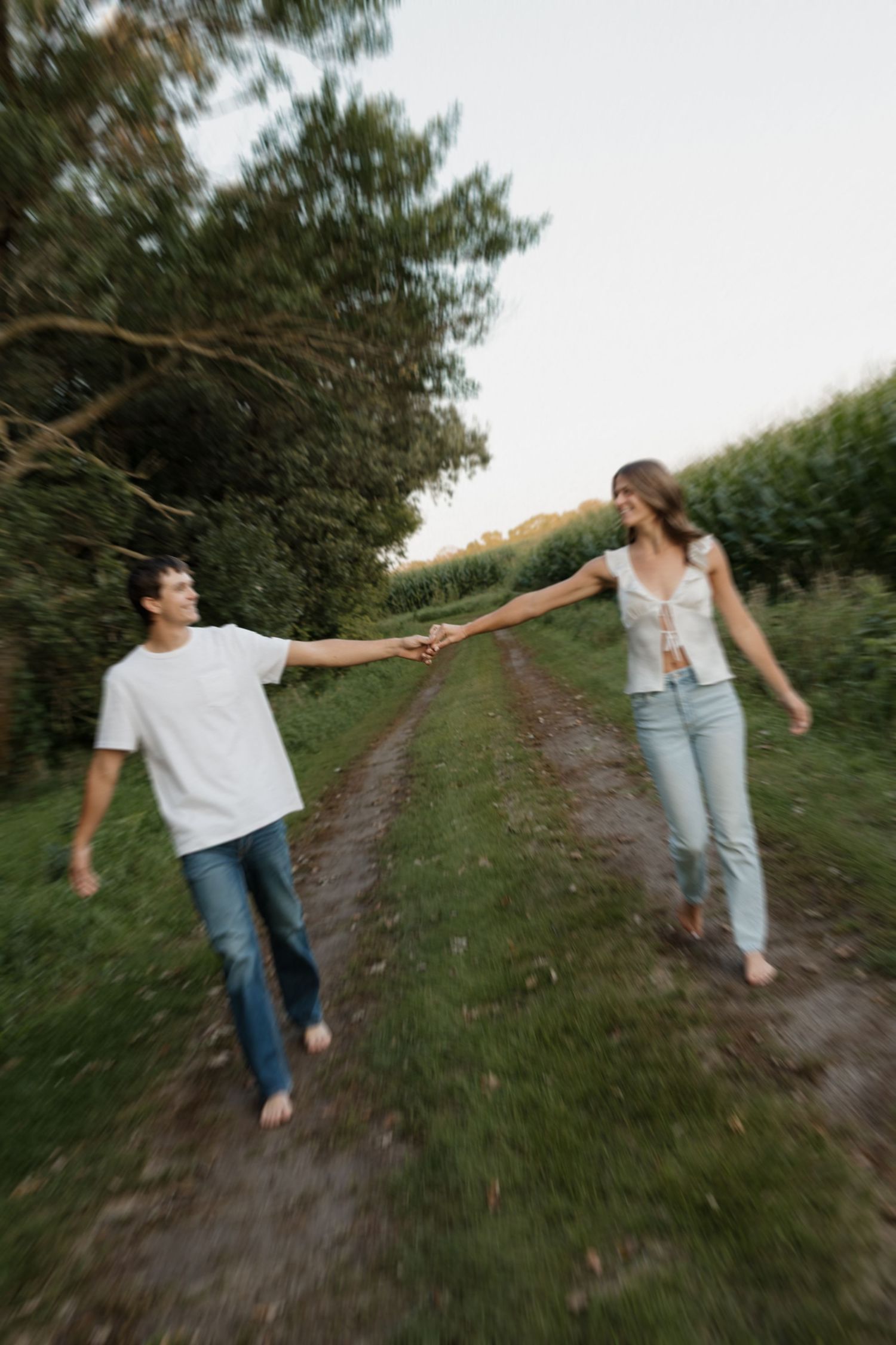 Playful couple holding hands and walking along a rural dirt path surrounded by lush greenery.