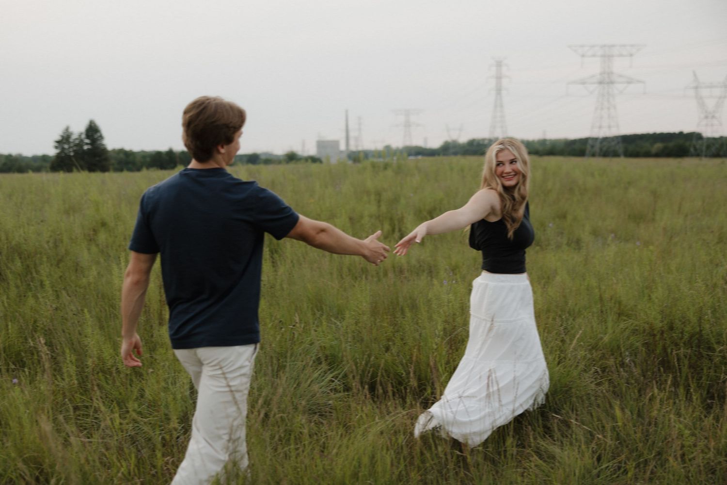 Playful couple spinning and holding hands in a wide green meadow with power lines in the distance.