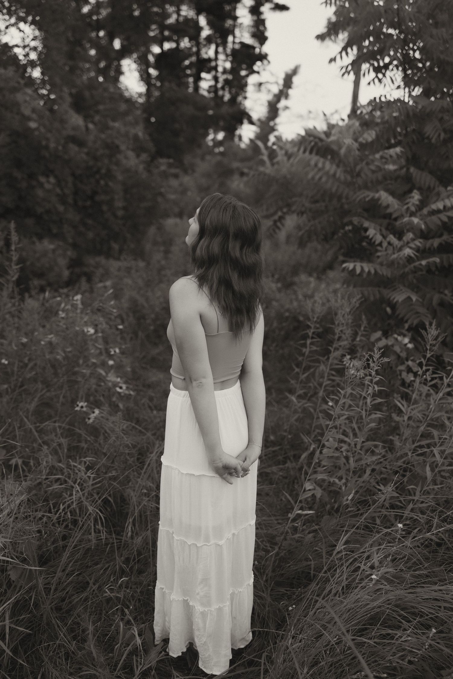 Black and white photo of a young woman in a backless white dress standing in a lush forest.