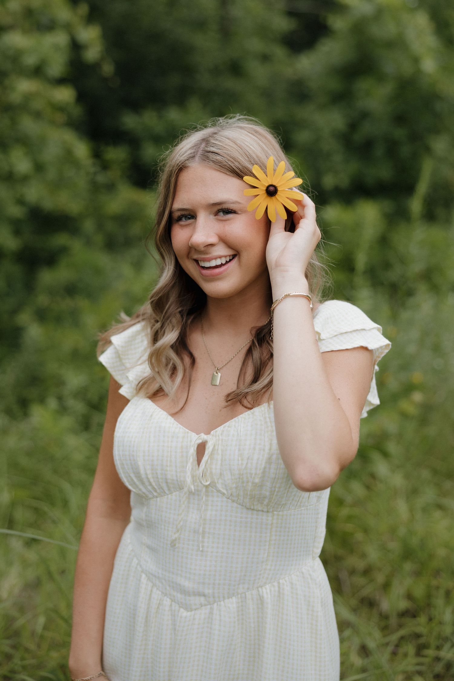 Smiling blonde woman in white dress holds a yellow sunflower near her face in green field.