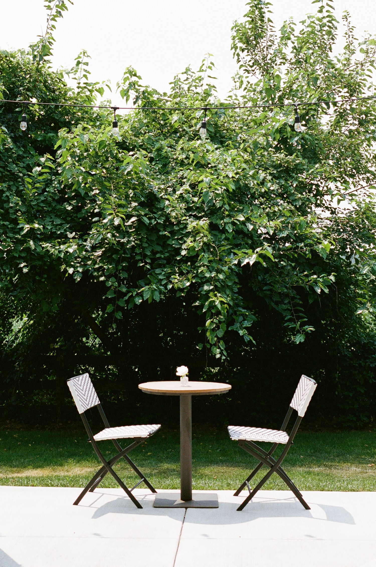 Two folding chairs and a round bistro table on a patio surrounded by lush green trees.