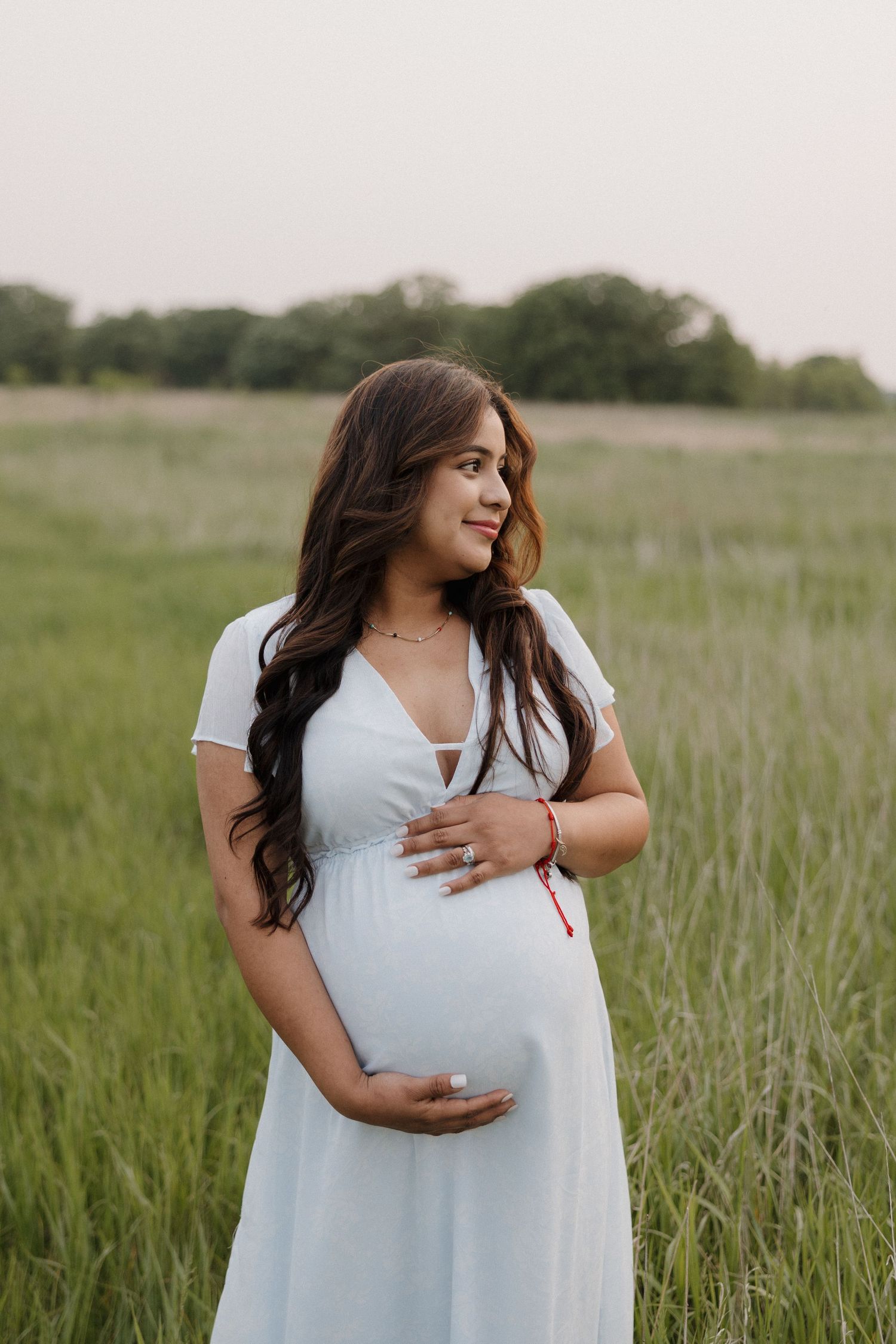 Pregnant woman in white maxi dress standing in green field cradling baby bump at sunset.