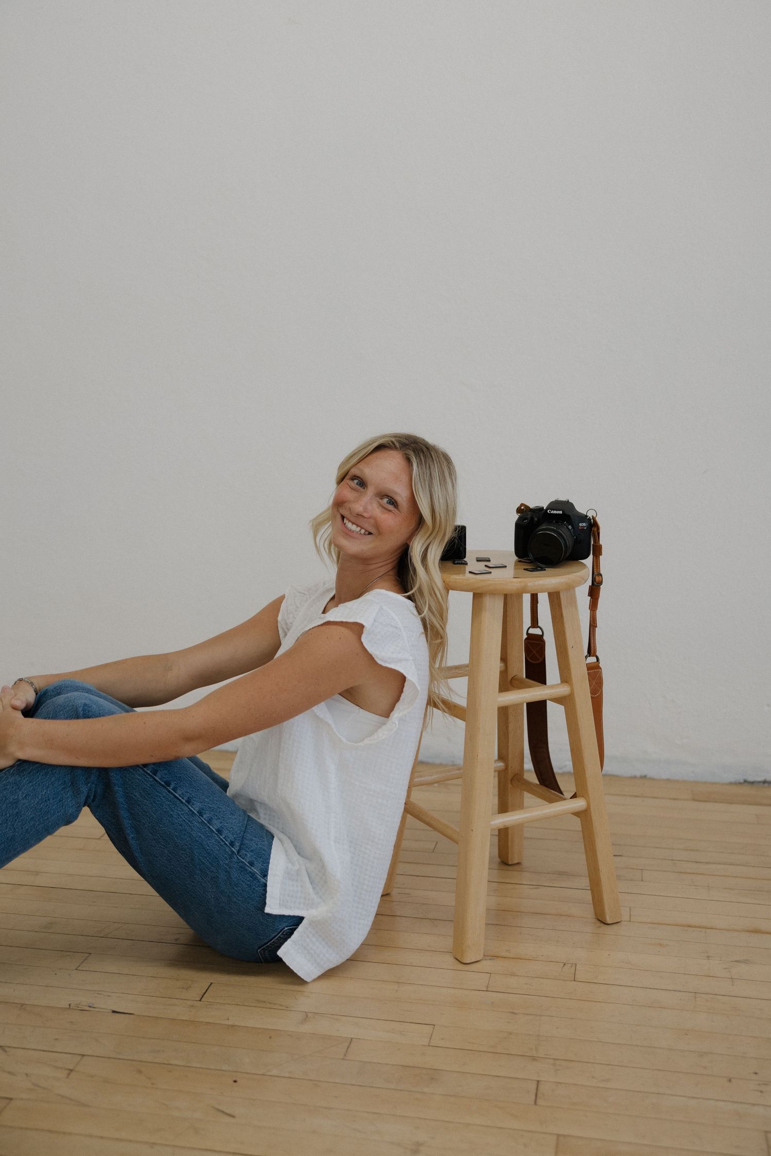 Smiling blonde woman sitting on floor beside a wooden stool with a camera in a studio.