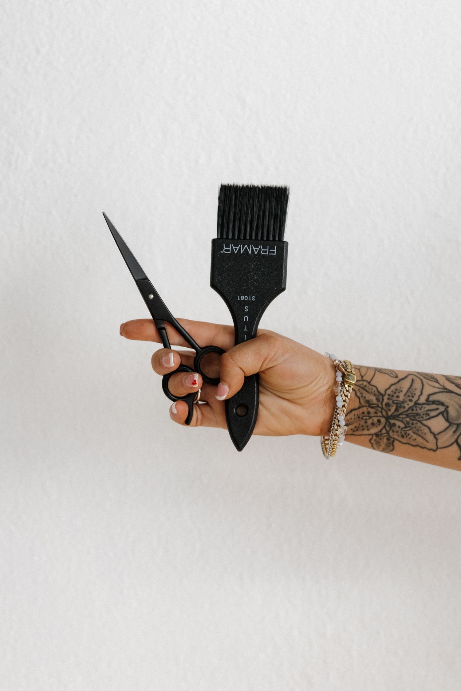 Tattooed hand holding black Framar hair dye brush and scissors against white wall.