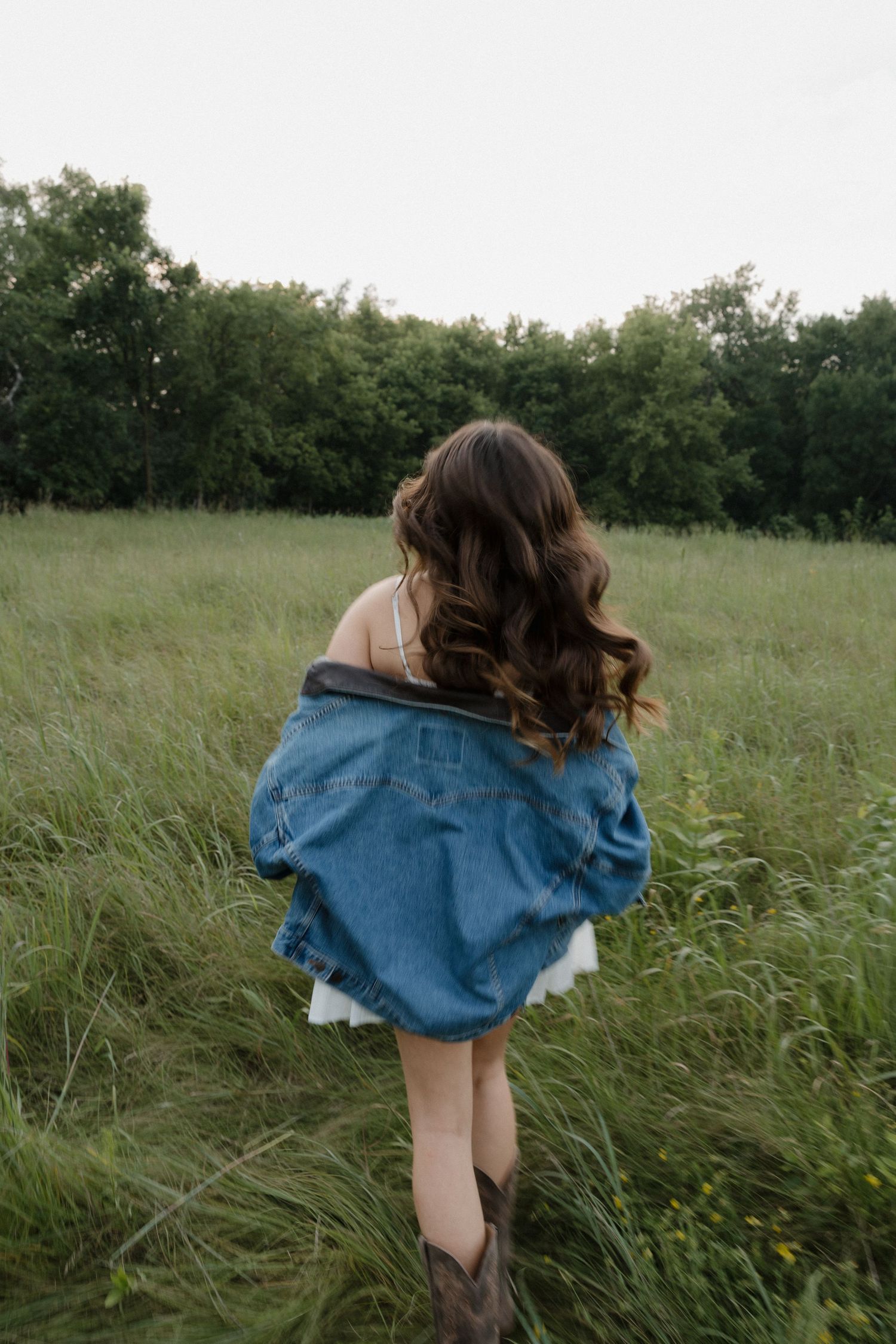 Young woman with long wavy hair in denim jacket and white dress walking through tall green grass field.