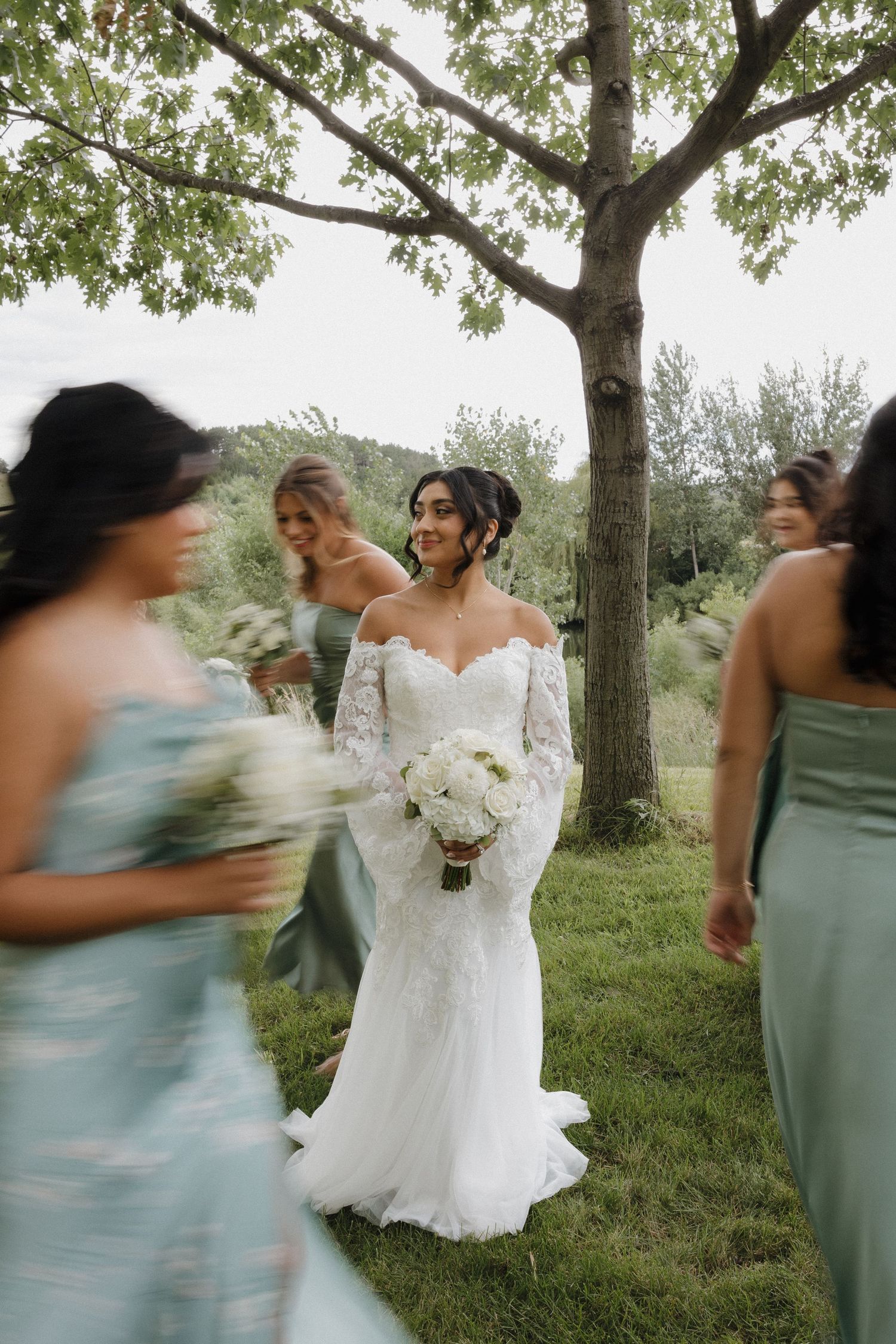 A bride in a white lace gown shares joyful moments with bridesmaids in sage green dresses under tree canopy.
