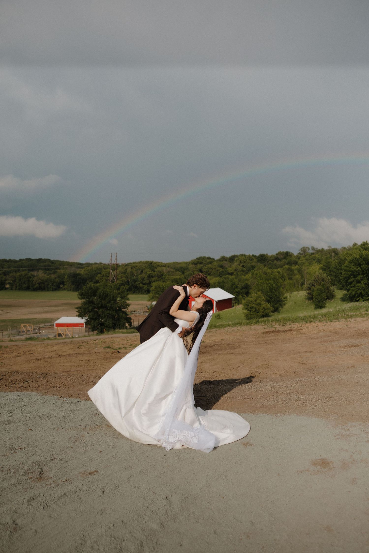 A white wedding dress flows gracefully across sandy ground against a cloudy sky with a rainbow in the background.