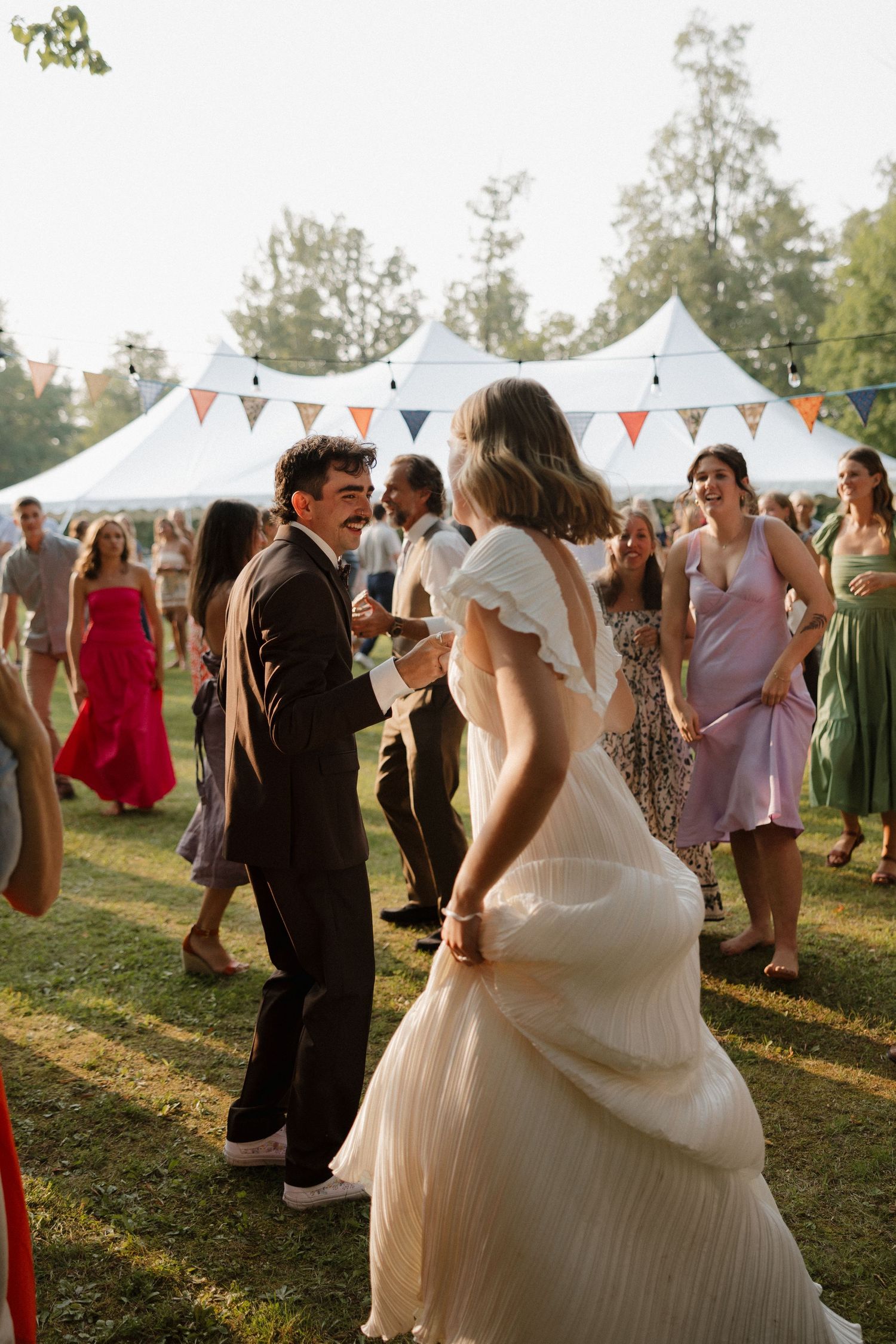 Wedding celebration outdoors with white tents and guests dancing in the golden sunset light.
