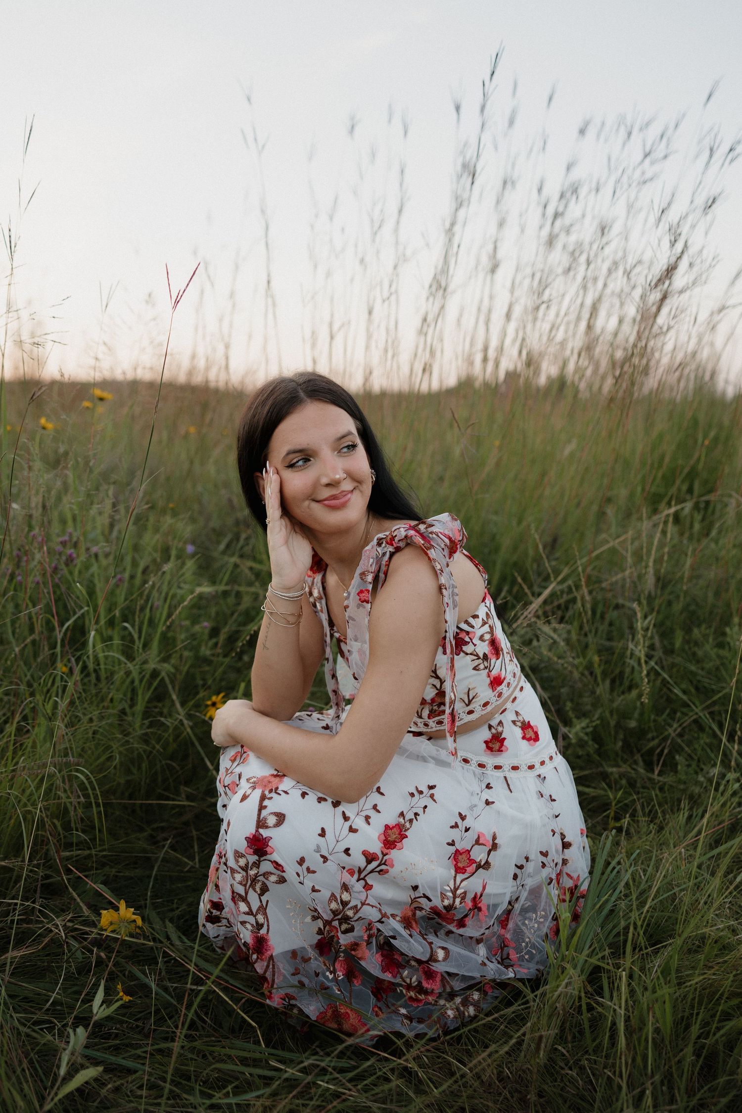 Dark-haired young woman in floral dress sitting gracefully in tall green grass at golden hour.