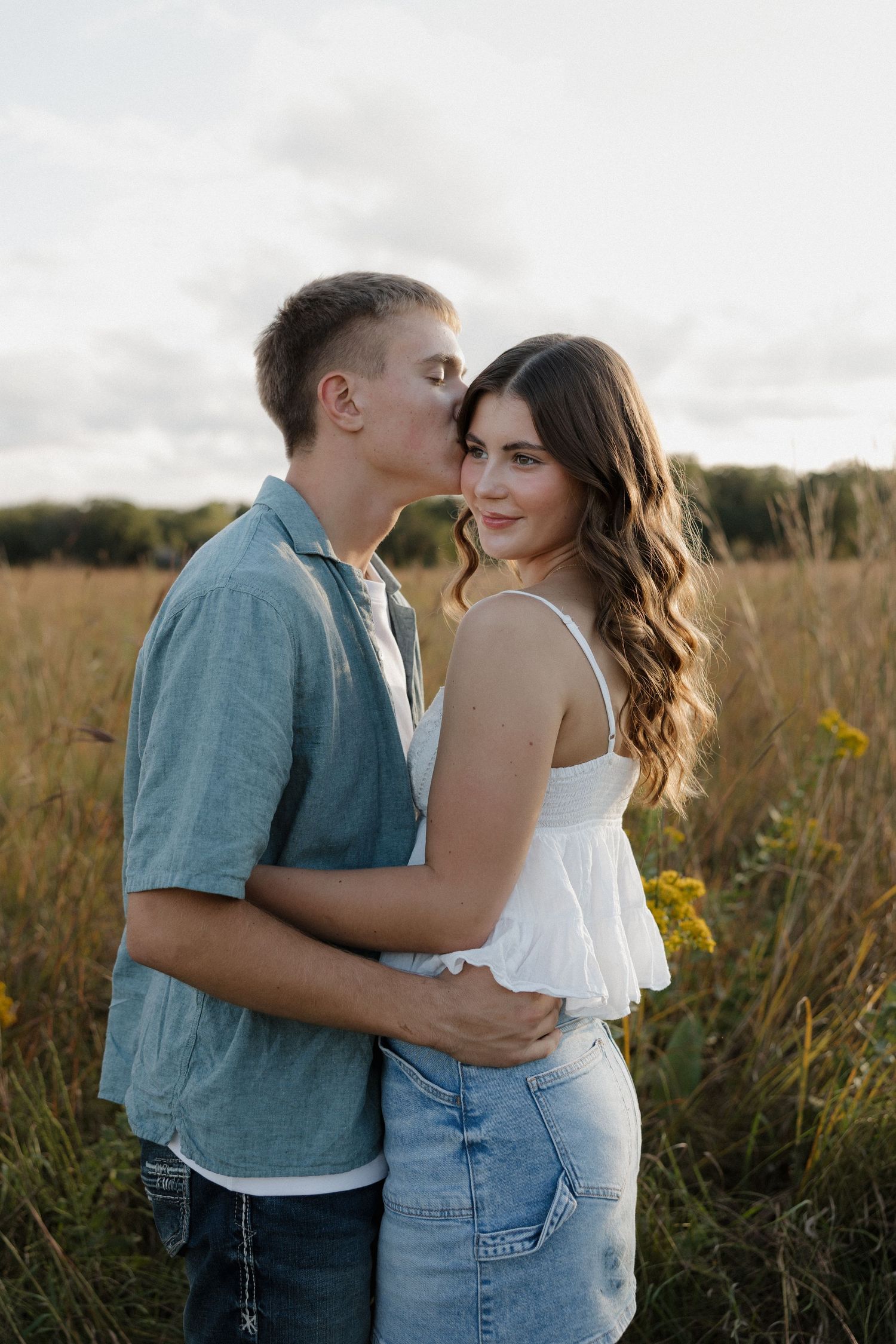 Young couple embracing in golden field, man kissing woman's cheek as she smiles at camera.
