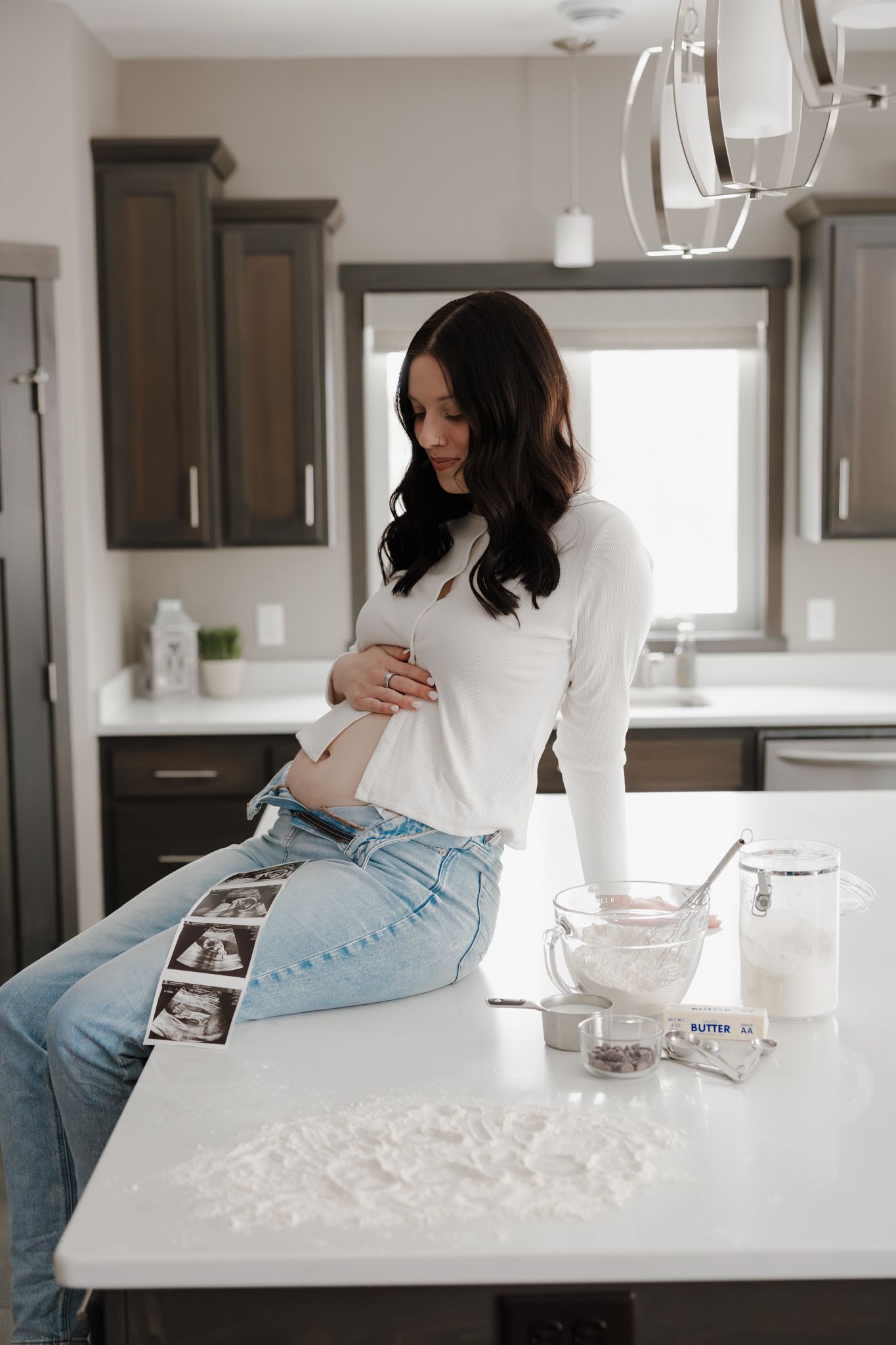 Pregnant woman in white top and jeans sitting on kitchen counter with ultrasound photos nearby.