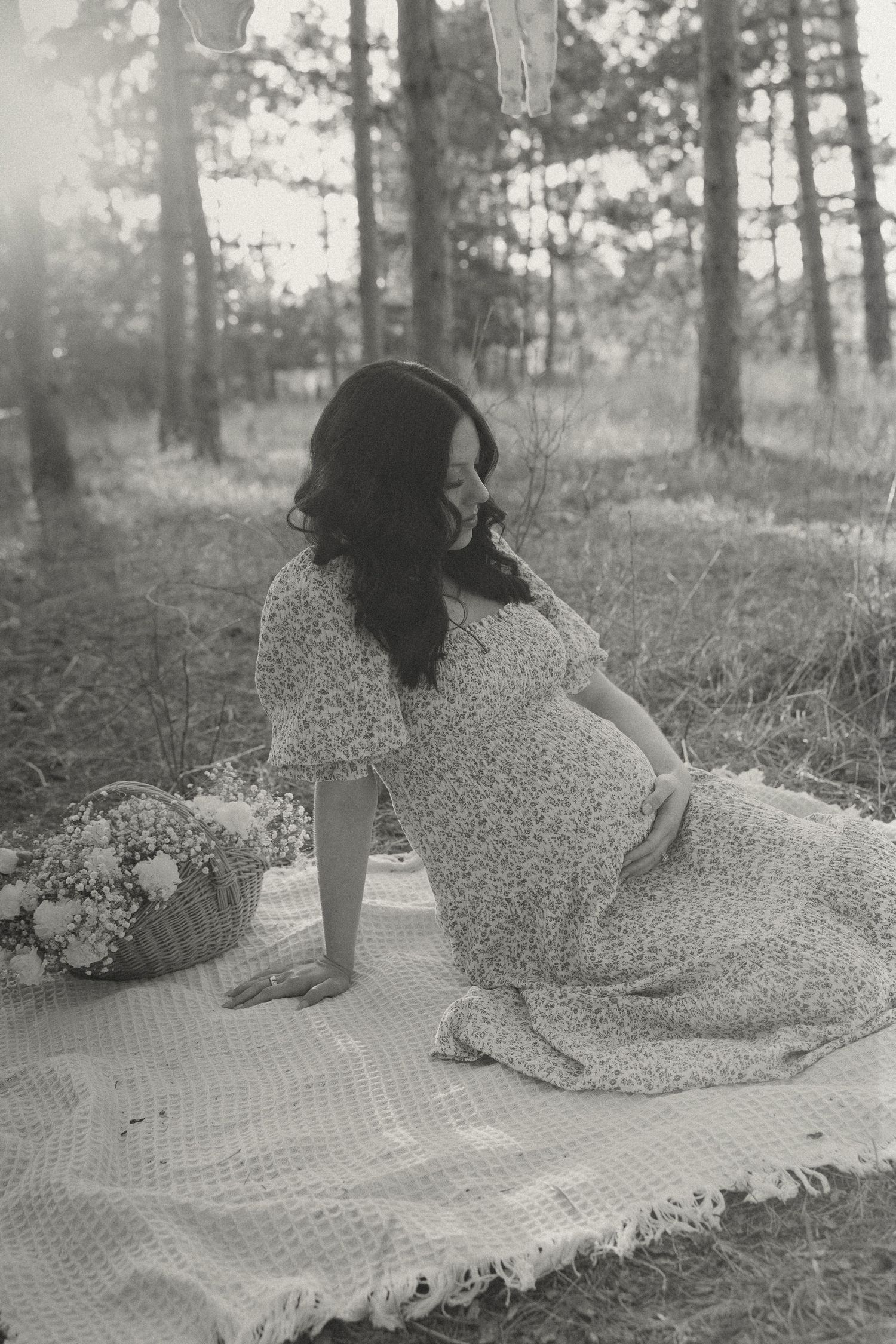 Black and white maternity photo of pregnant woman in floral dress sitting on blanket in forest.