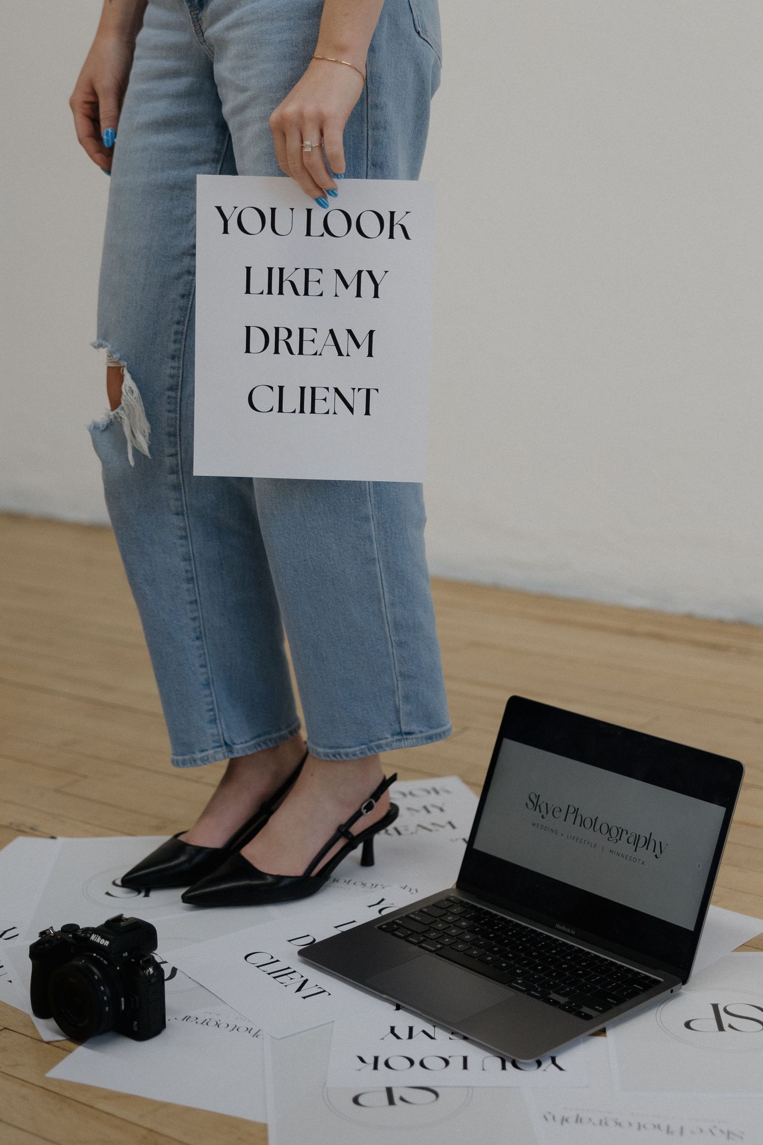 Photographer holding 'You Look Like My Dream Client' sign with laptop and camera on studio floor.
