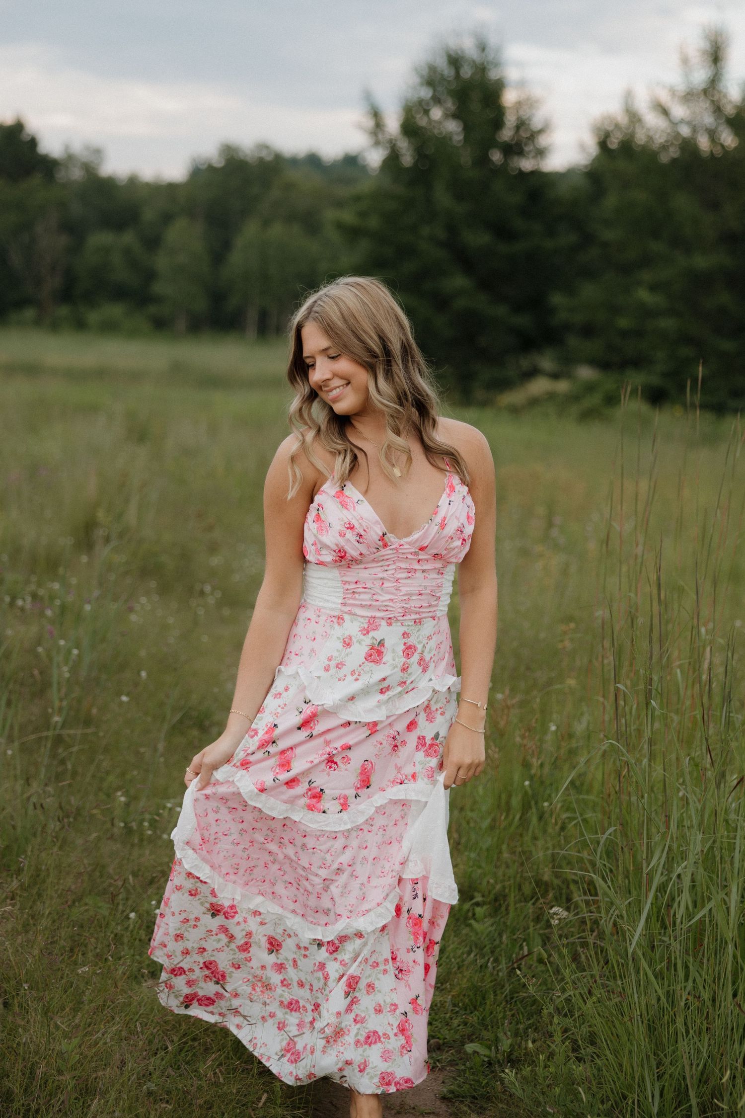 Smiling blonde woman in pink floral tiered maxi dress walks through a lush green meadow.