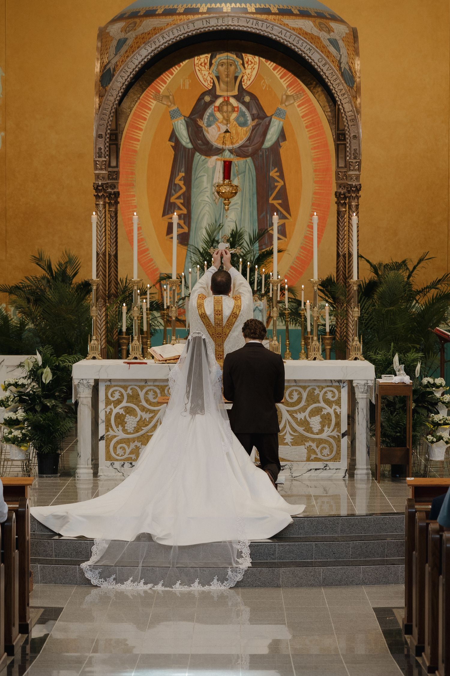 Wedding ceremony in ornate church altar with couple kneeling while bride's long white cathedral veil trails behind.