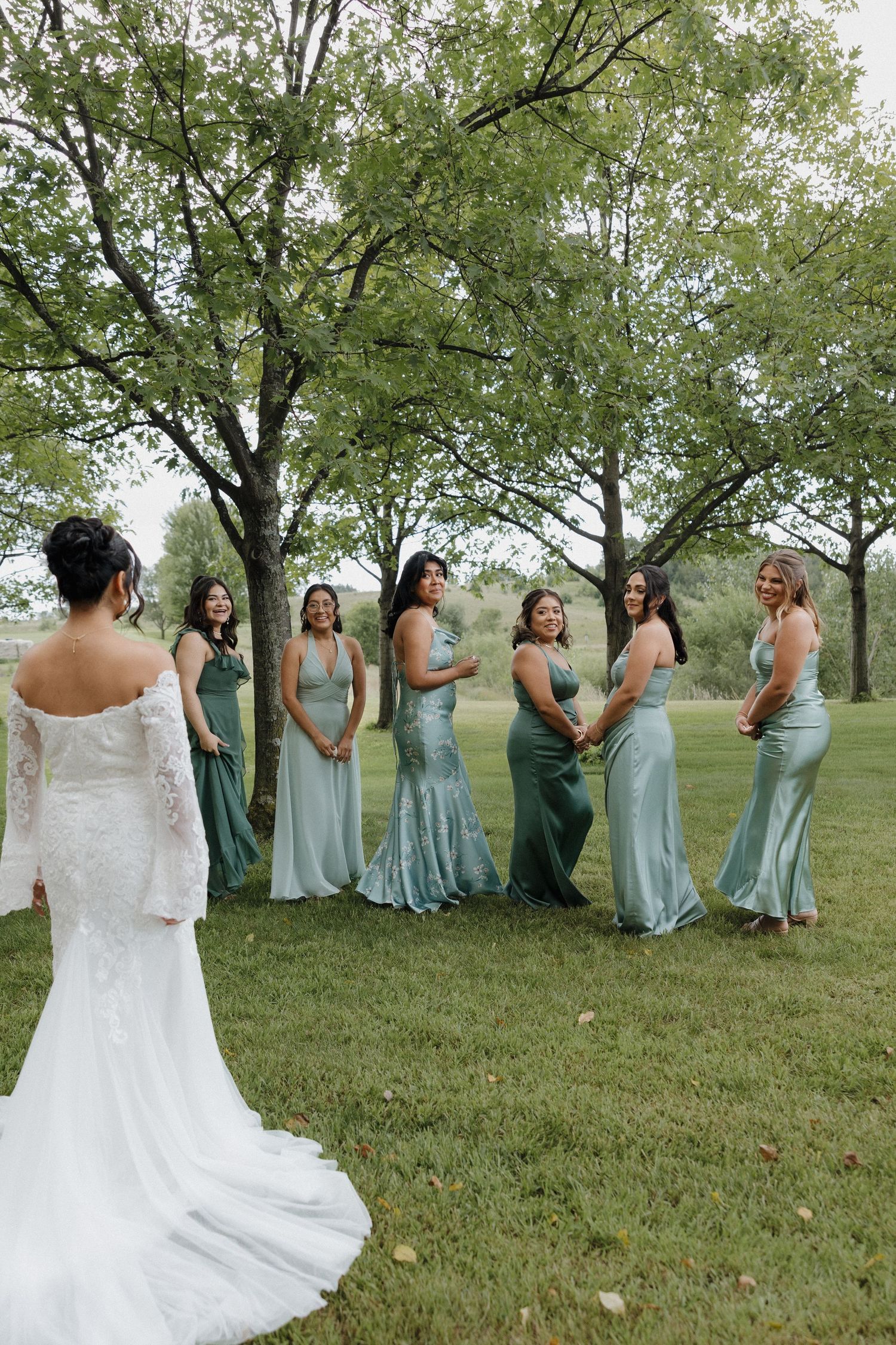Bridal party in mint green dresses posing outdoors under trees on grassy hill.