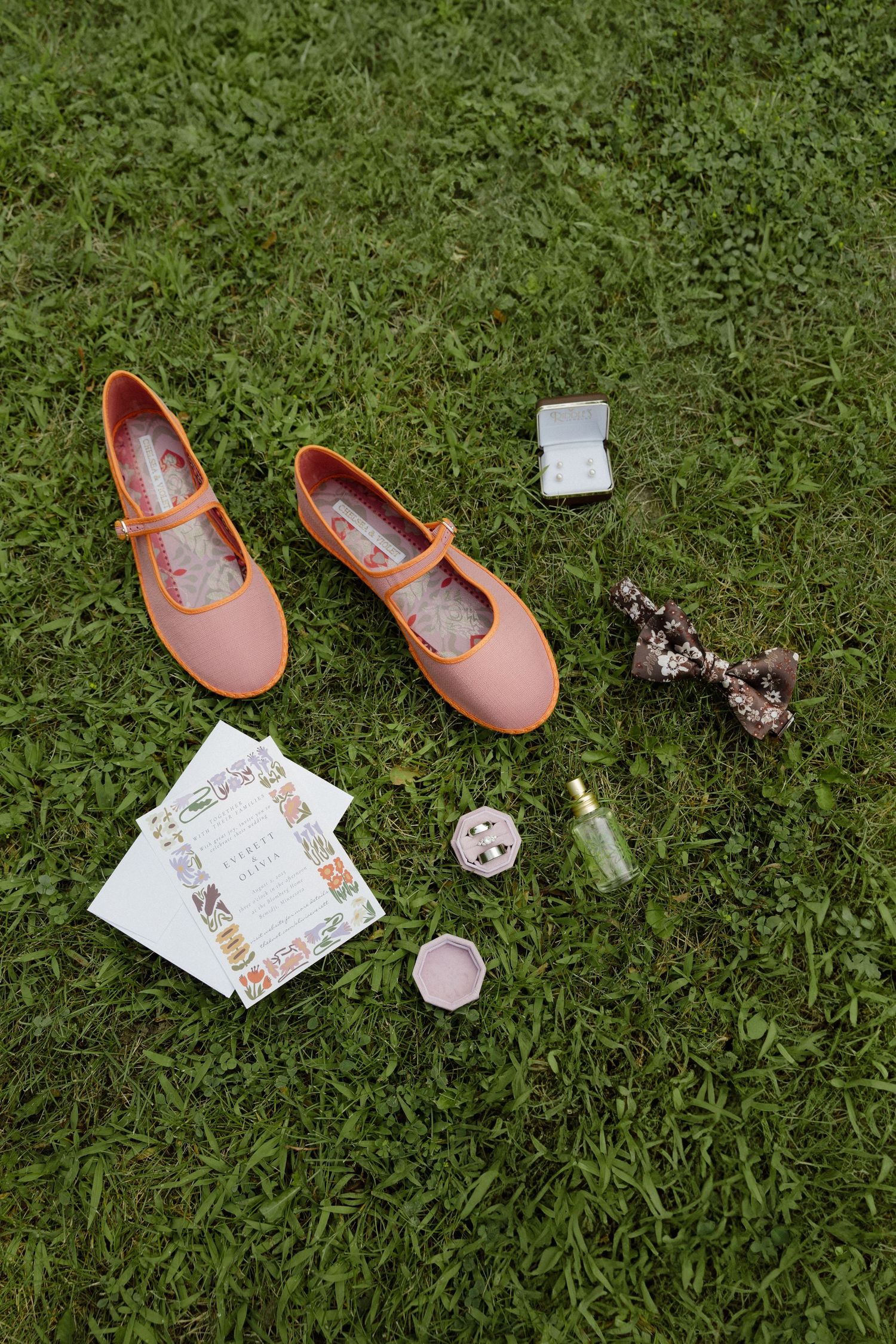 Pink ballet flats and wedding invitation details arranged on green grass.