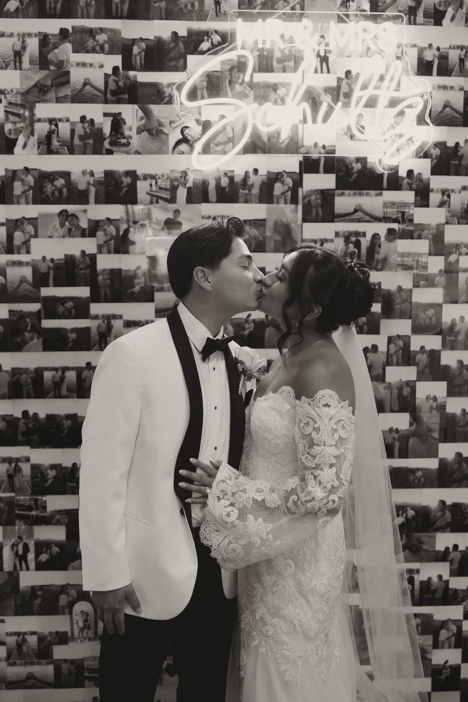 A romantic black and white wedding photo of a couple kissing in front of a wall covered with vintage photographs.