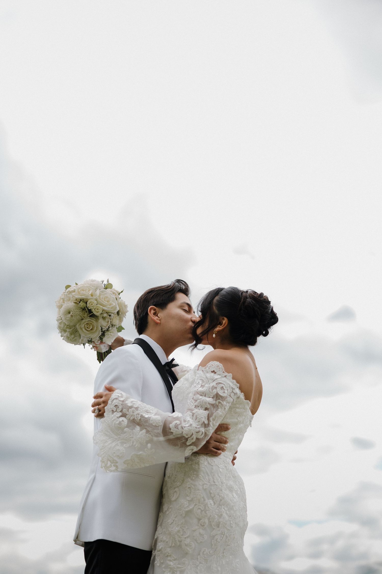 Bride and groom sharing a kiss under cloudy skies, bride in off-shoulder lace gown holding white roses.