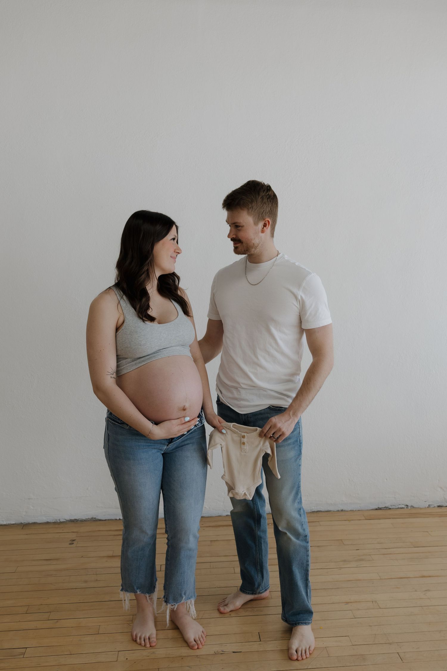 Pregnant woman and partner standing together holding baby onesie against white wall.
