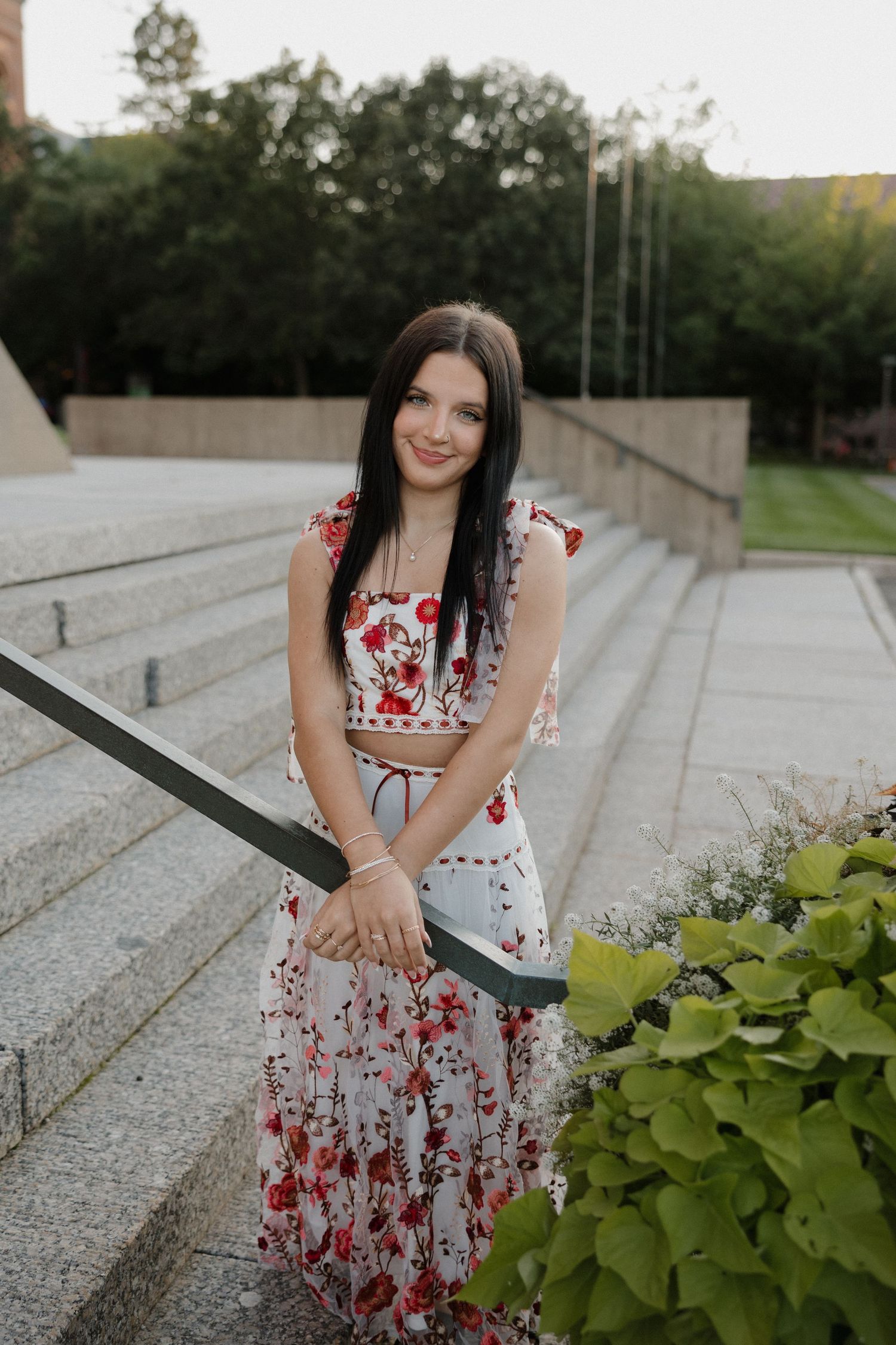 Dark-haired young woman in floral dress posing on stone steps near green plants at dusk.