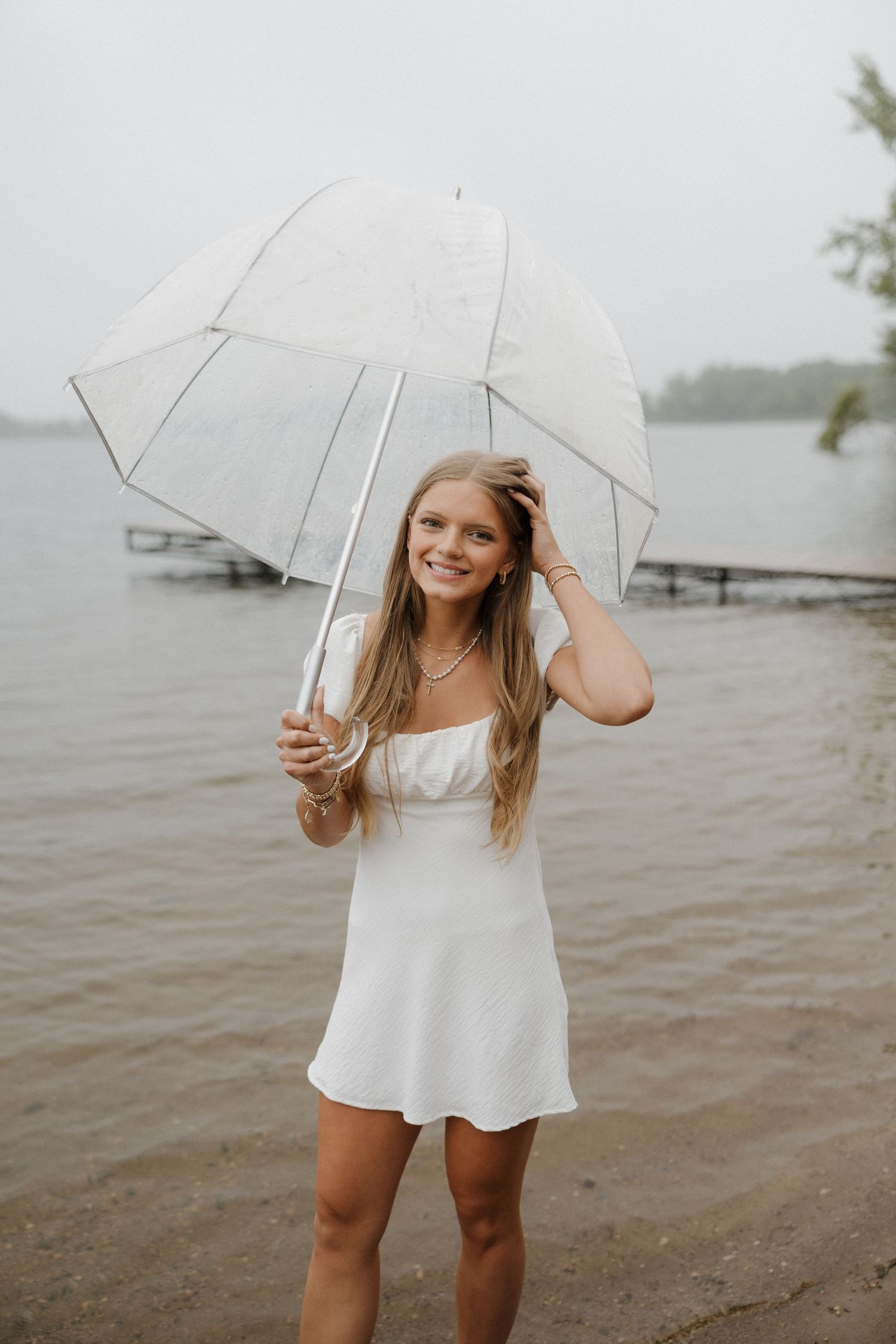 Smiling blonde teen in white mini dress holding a white umbrella on a rainy lakeside beach.