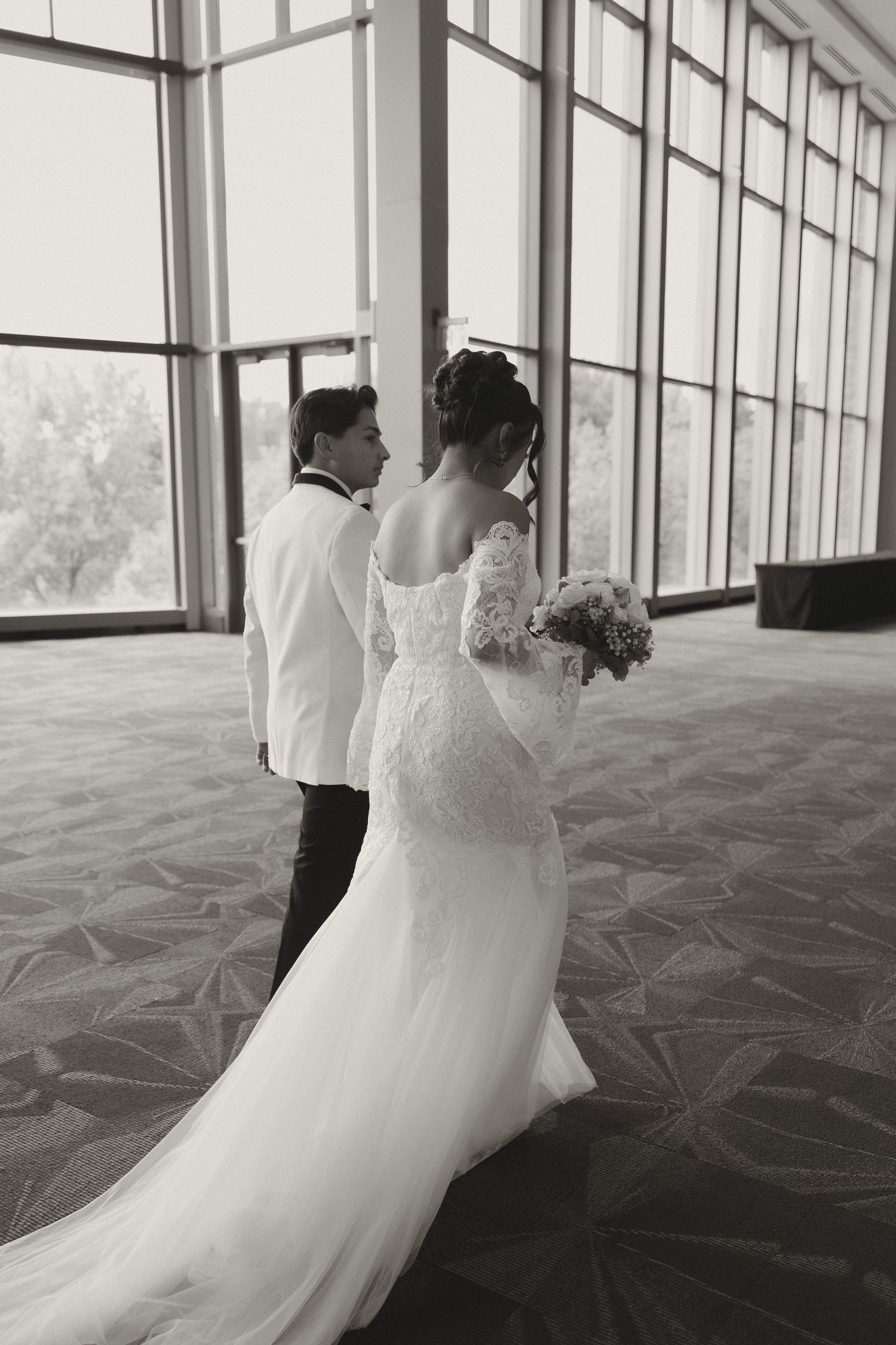Black and white photo of couple in wedding attire by large windows in modern venue.