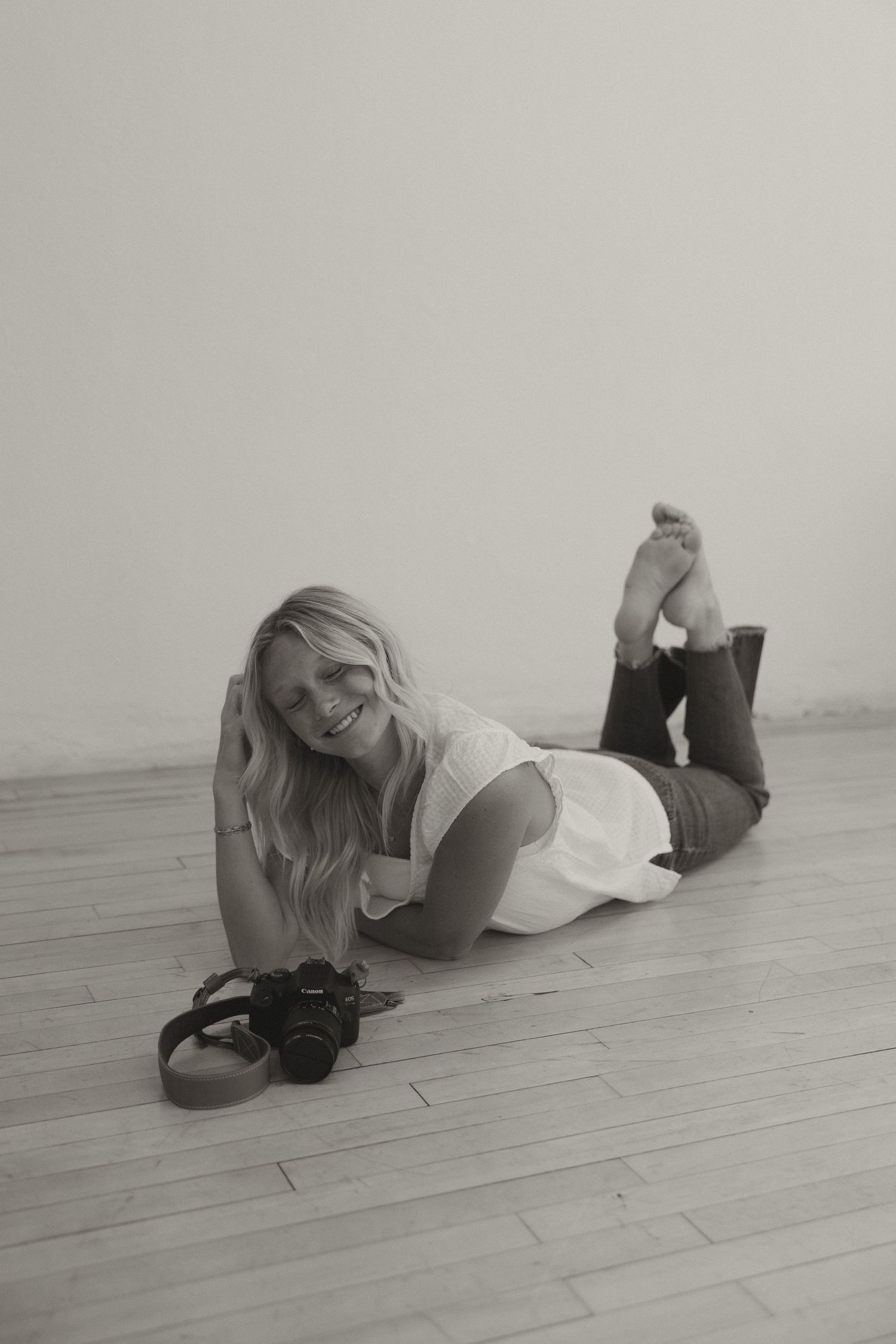 Black and white photo of a smiling blonde woman lying on a wooden floor with a camera.