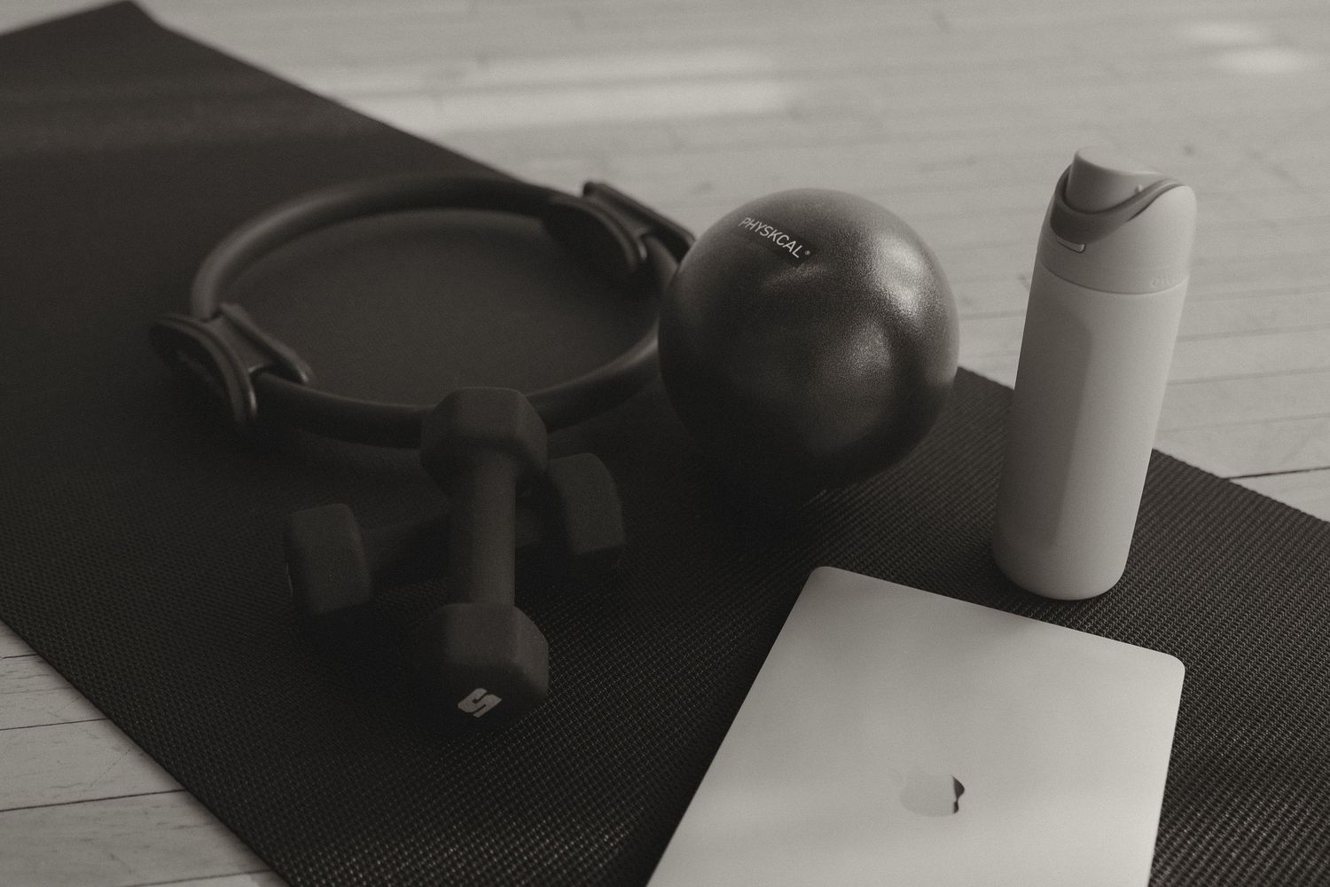 Black yoga mat with dumbbells, pilates ring, exercise ball, water bottle, and laptop.