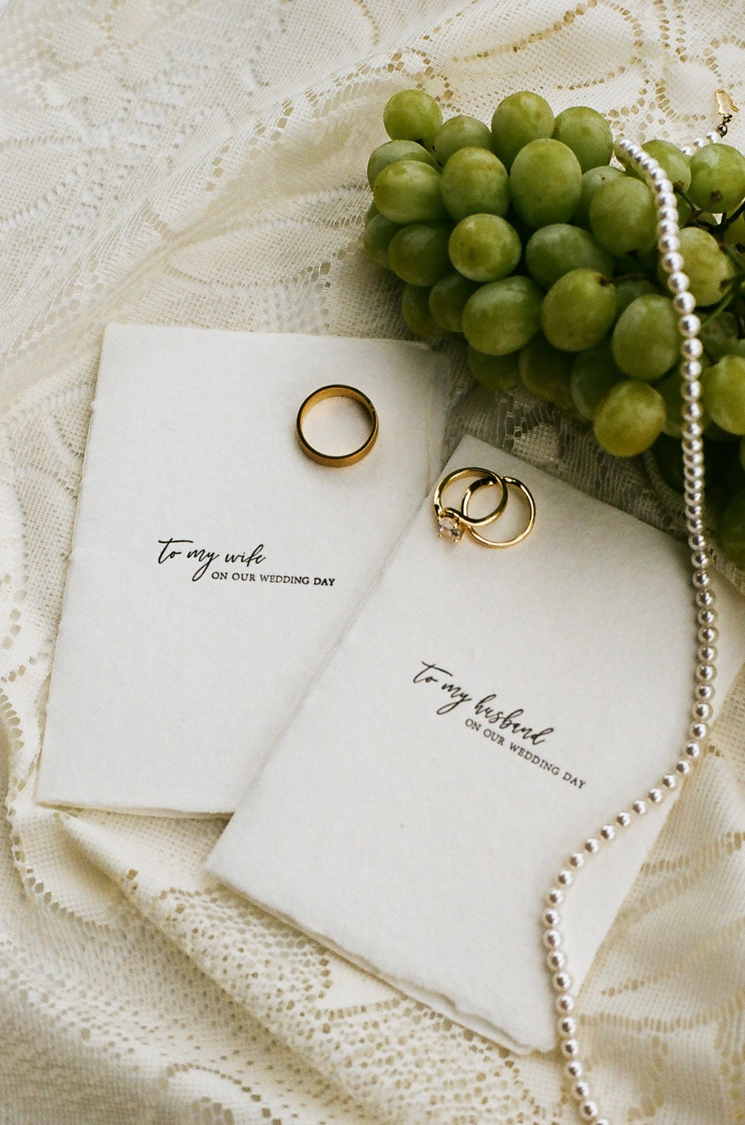 Wedding flat lay with gold rings, pearl necklace, green grapes, and cards reading to my wife and to my husband.