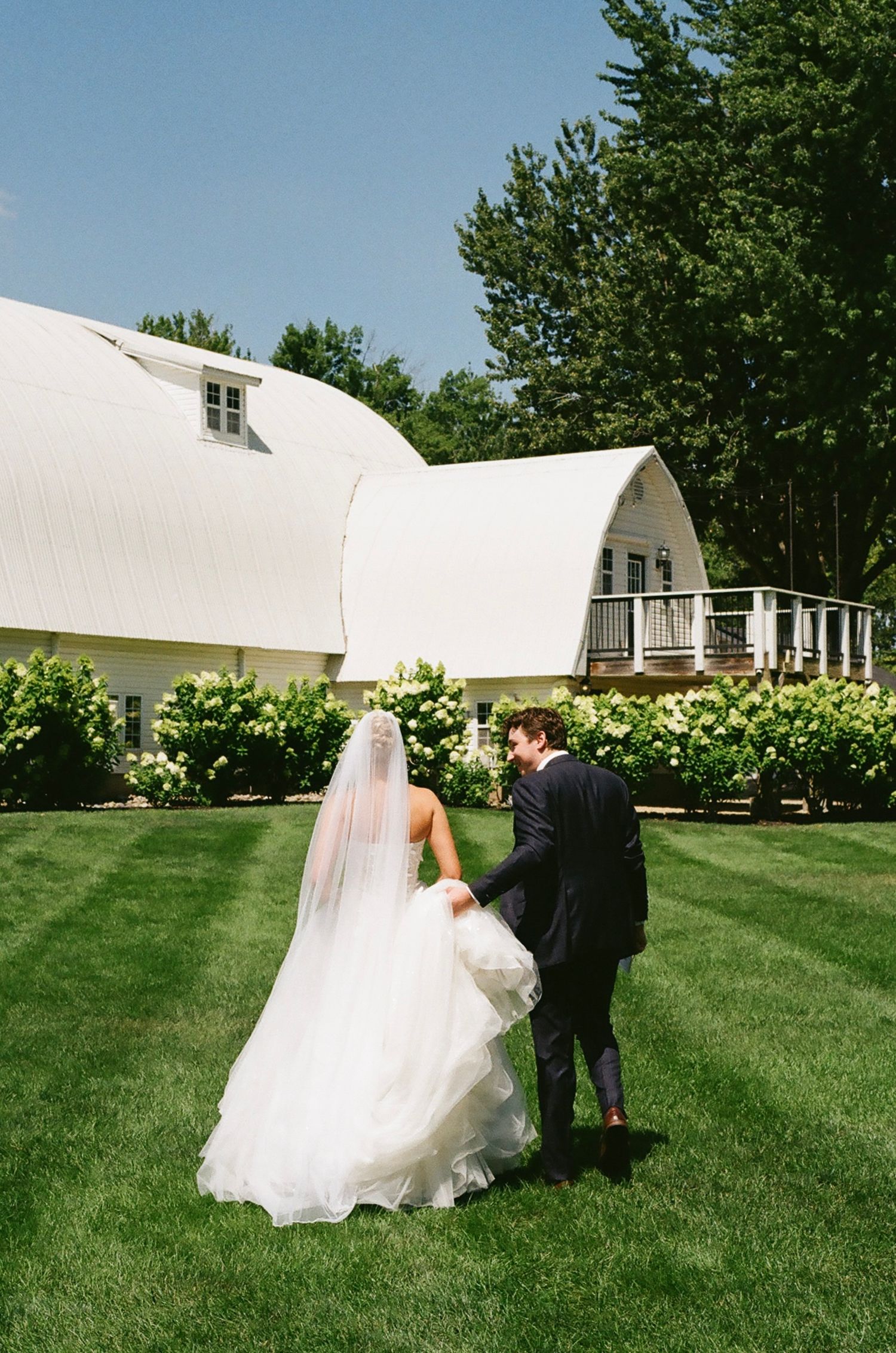 Bride and groom walk hand-in-hand across lush green lawn toward white barn venue surrounded by trees.