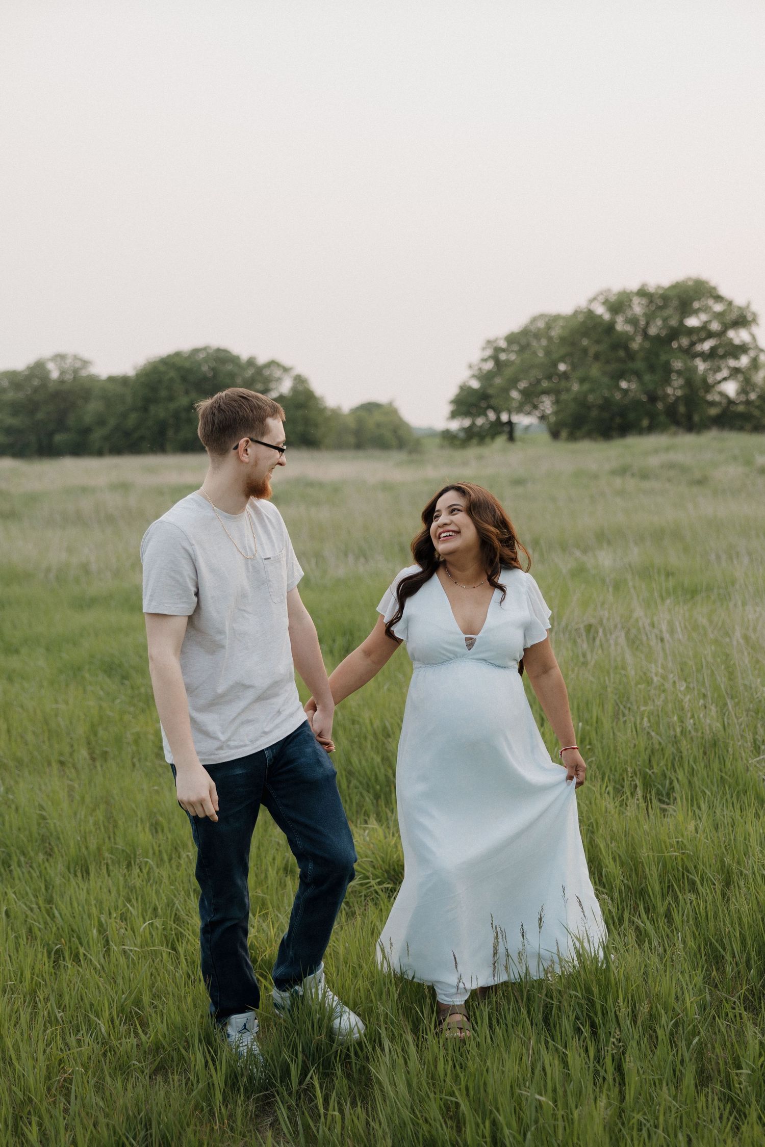 Pregnant woman in light blue maxi dress walking hand in hand with partner through green meadow.