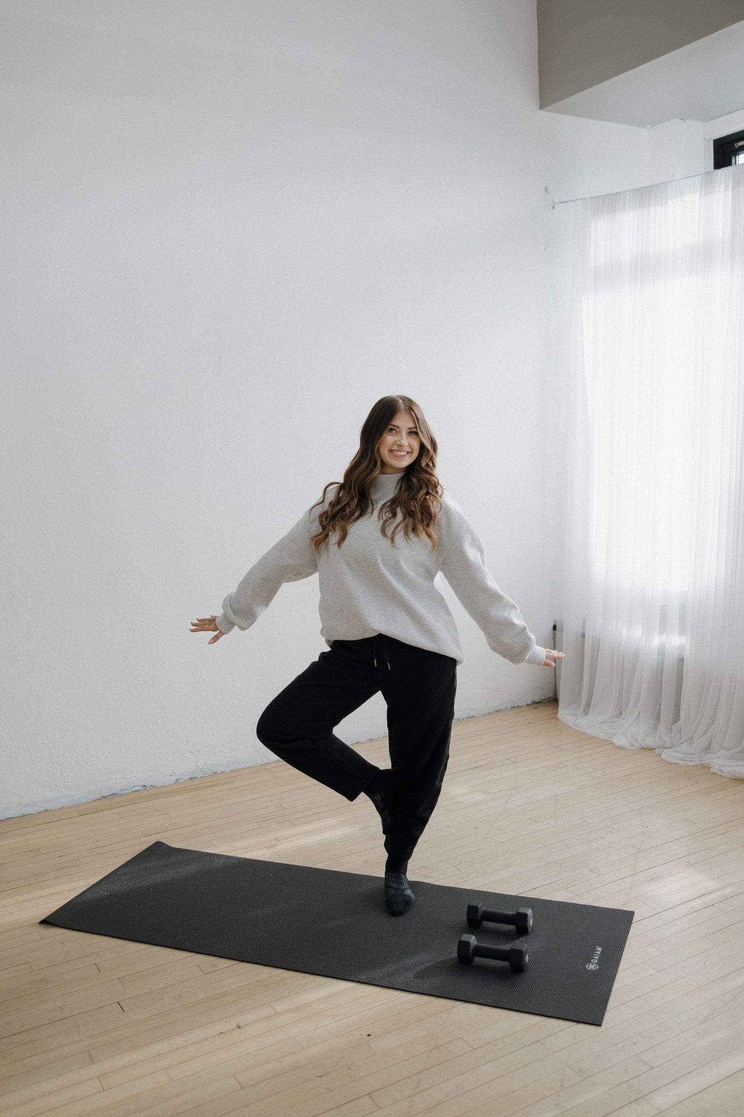 Smiling woman in gray sweater balancing in tree pose on a black yoga mat with dumbbells.