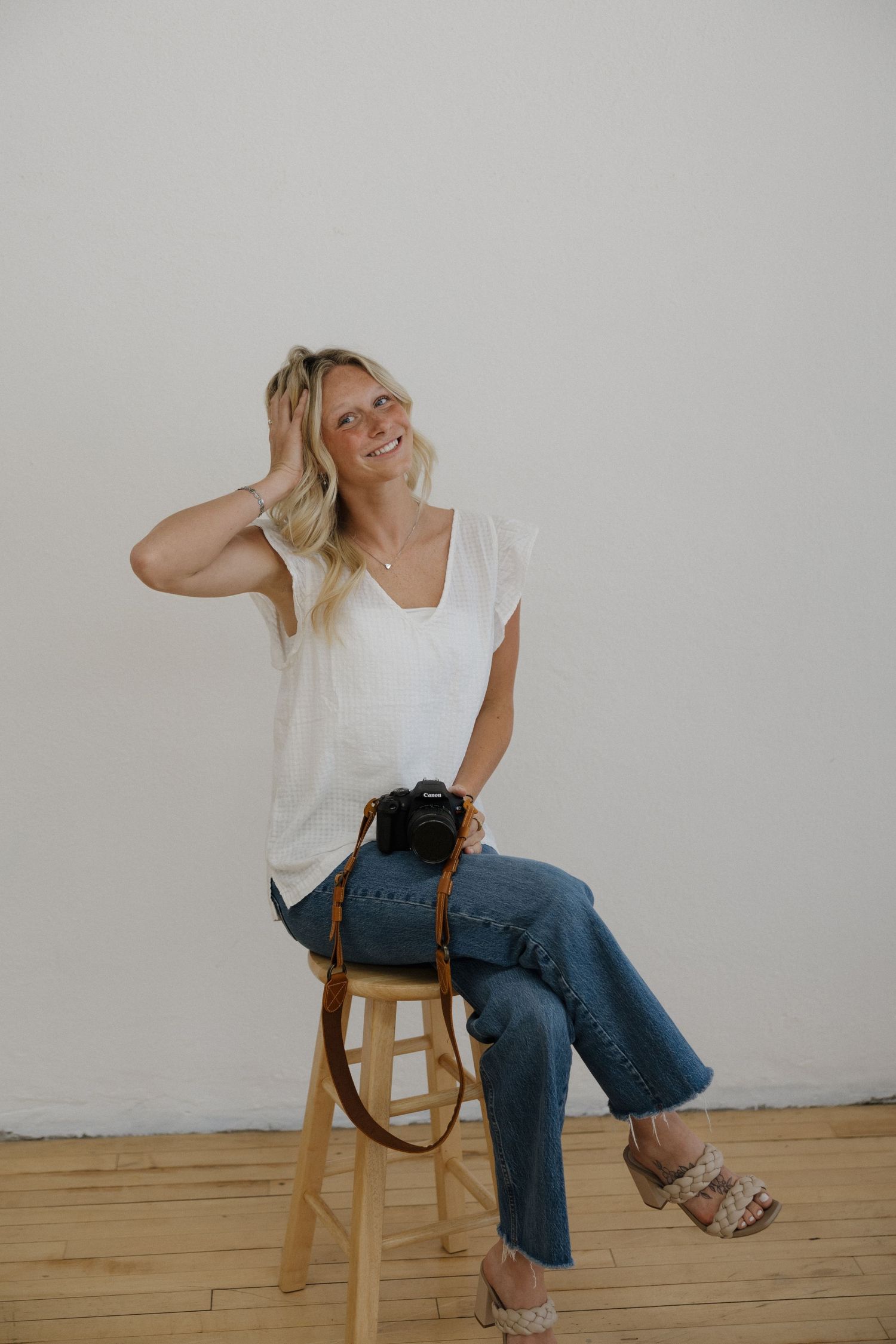 Smiling blonde woman sitting on a wooden stool holding a camera in a bright studio.