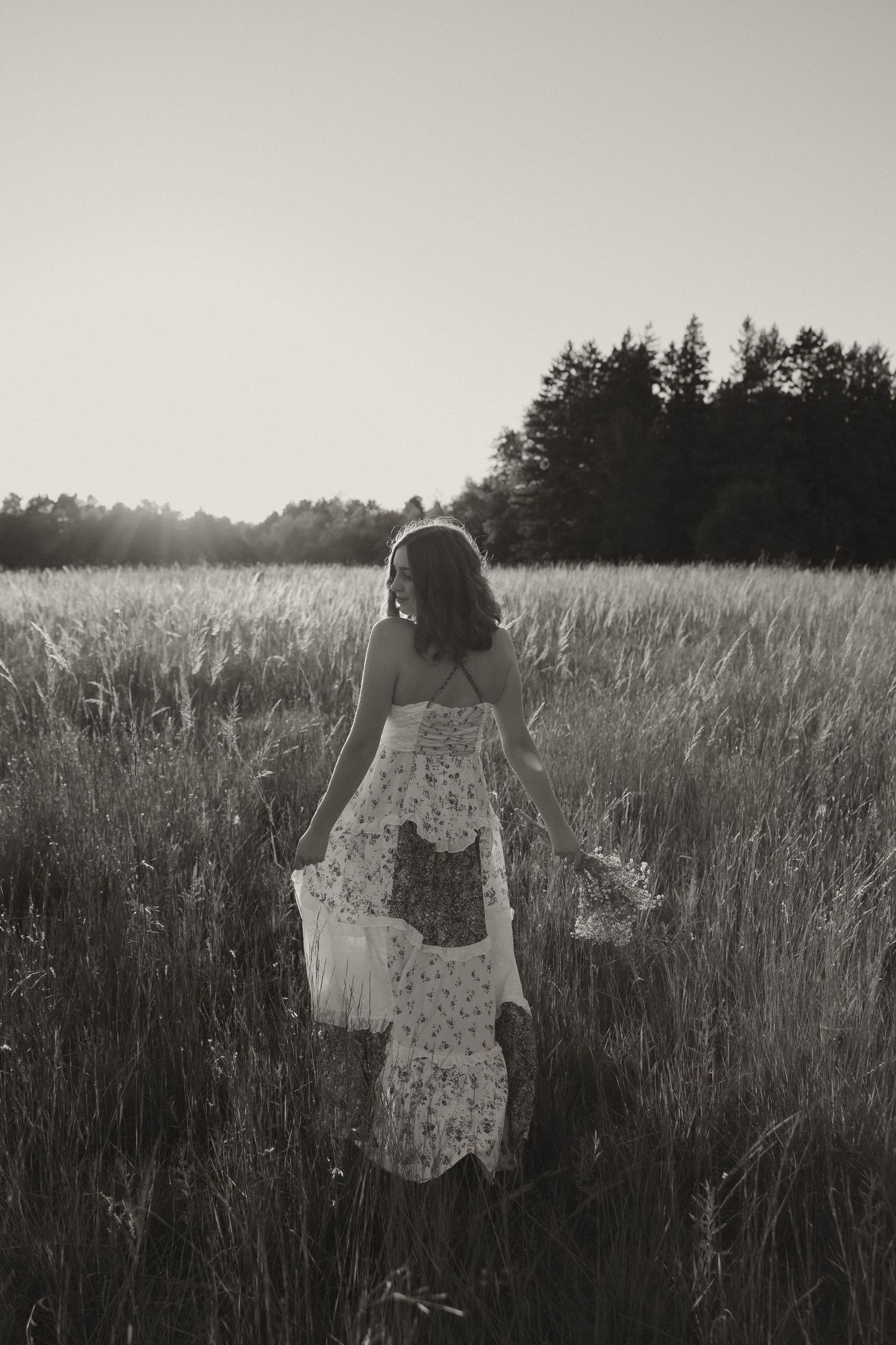 Black and white photo of woman in floral dress walking through golden field at sunset.