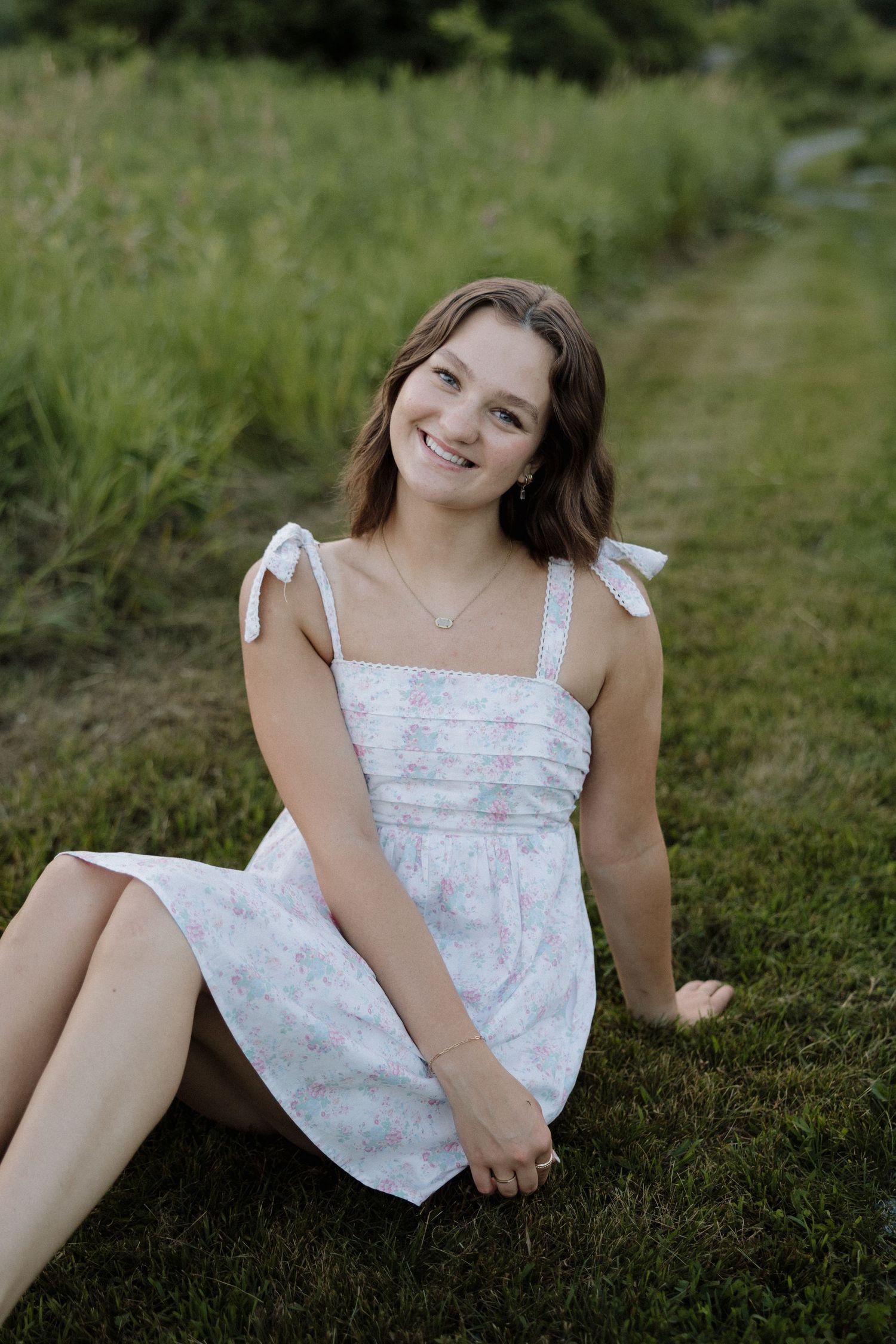 Smiling young woman in a floral sundress sitting on green grass near tall reeds outdoors.