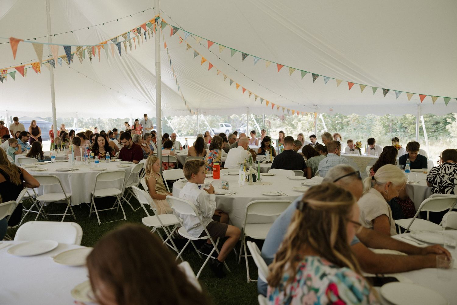 A large white tent filled with guests seated at tables during an outdoor celebration with colorful bunting decorations.