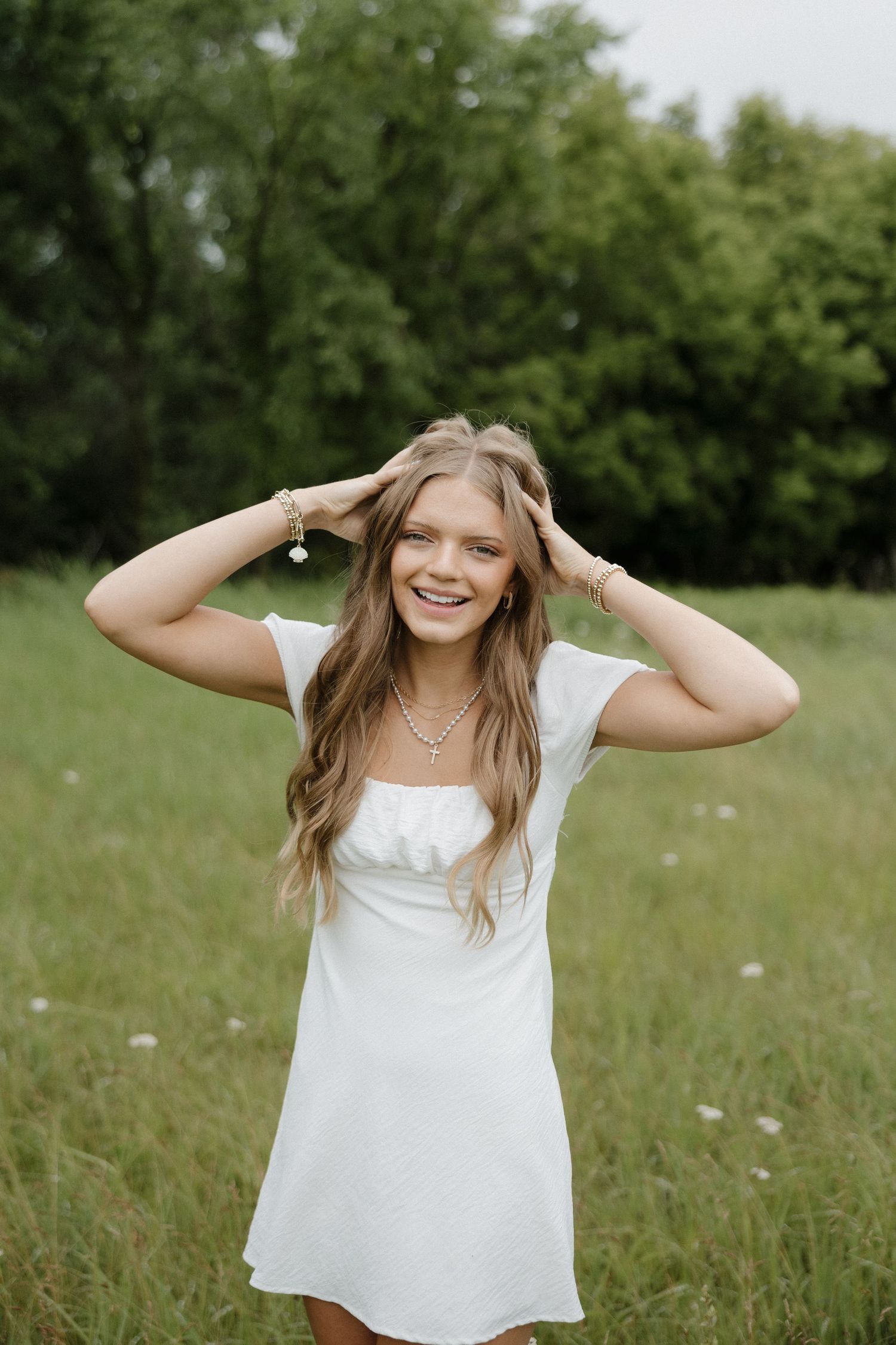 Smiling young woman in white mini dress posing in a green meadow with hands in hair.