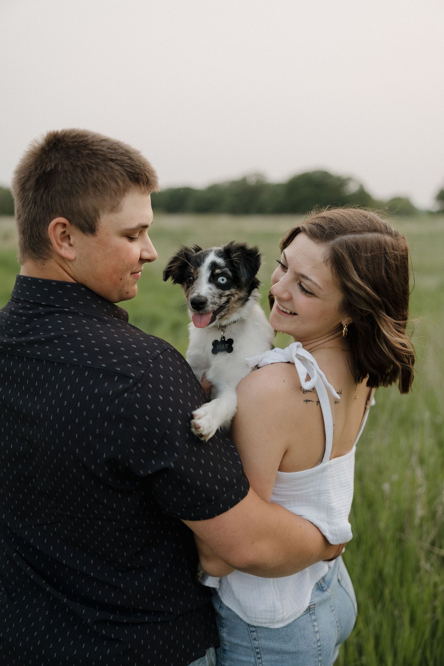 Happy couple holding an adorable black and white puppy together in a lush green field at sunset.