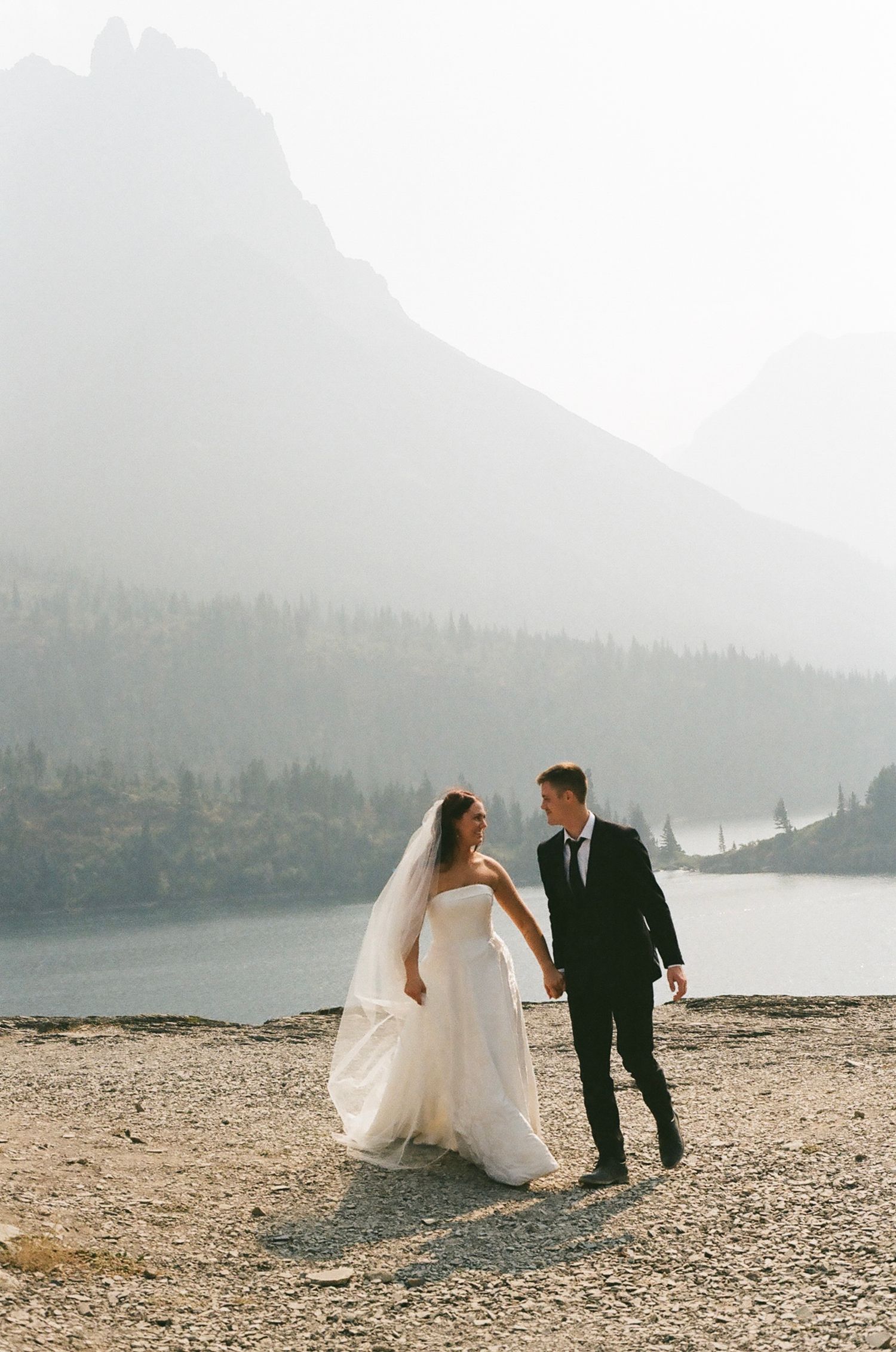 Bride and groom hold hands walking near a misty mountain lake, bride in white gown with veil, groom in black suit.