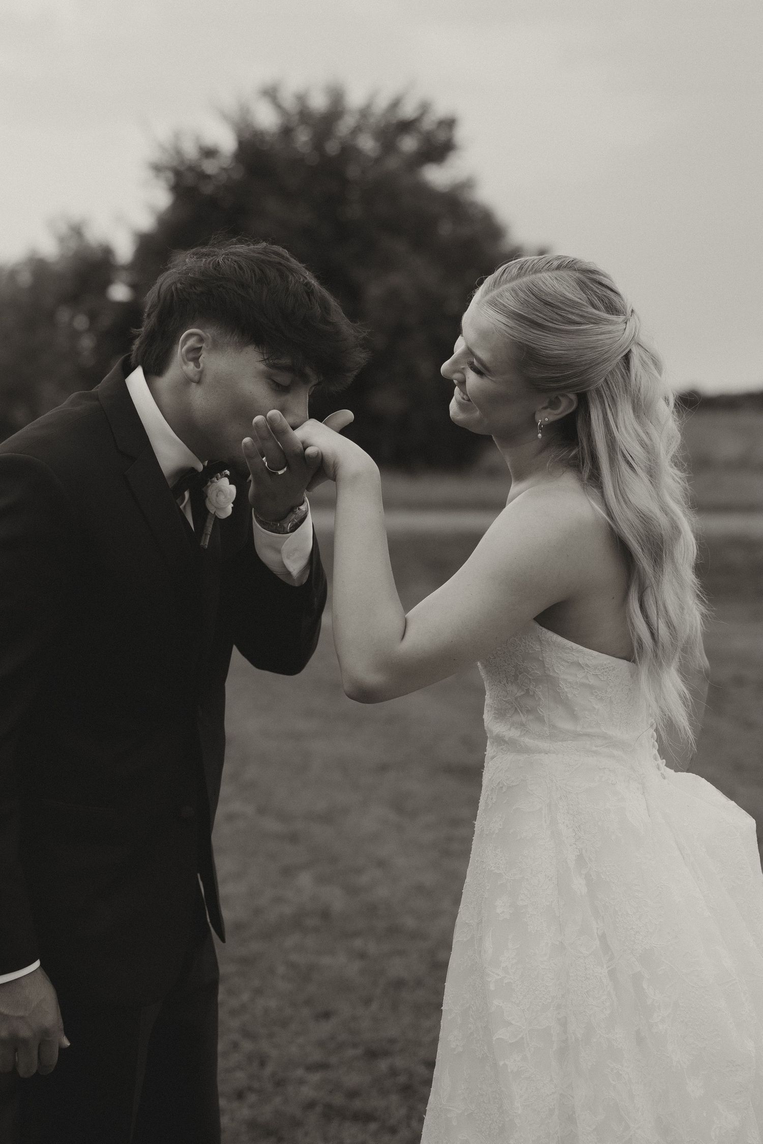 Black and white photo of a groom kissing his bride's hand on their wedding day outdoors.
