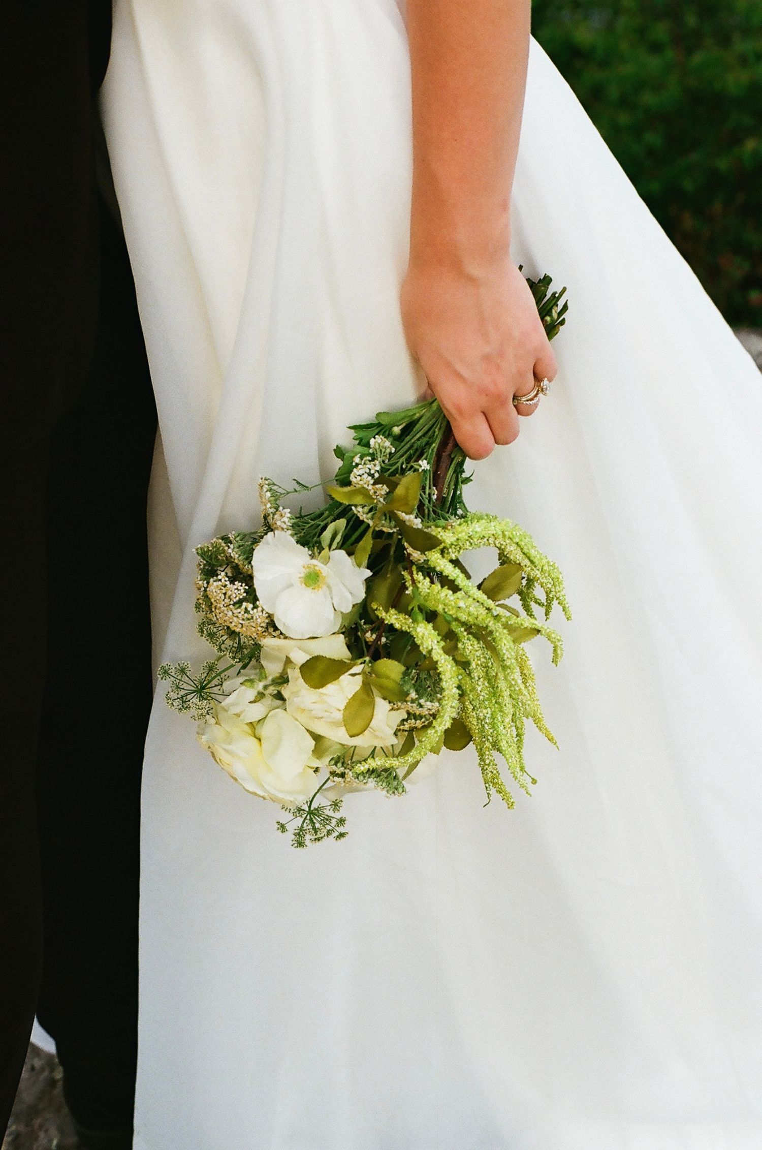 Bride holding a green and white wedding bouquet with roses and trailing amaranthus in her ringed hand against a white gown.